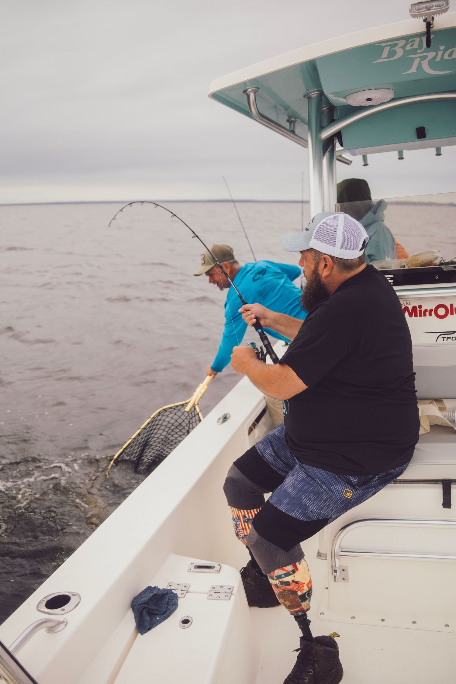 Two men fishing from a boat on the ocean, one reeling in a catch with a bent rod and net ready.