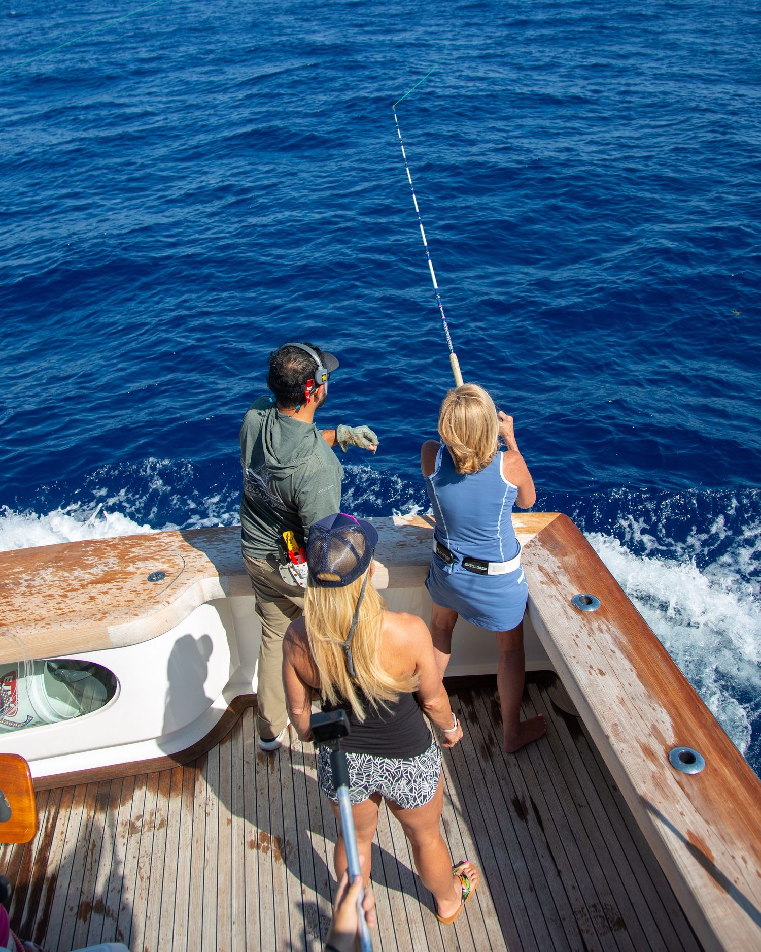 Three people fishing from the stern of a boat on deep blue ocean waters, with a fishing rod bent over the sea.
