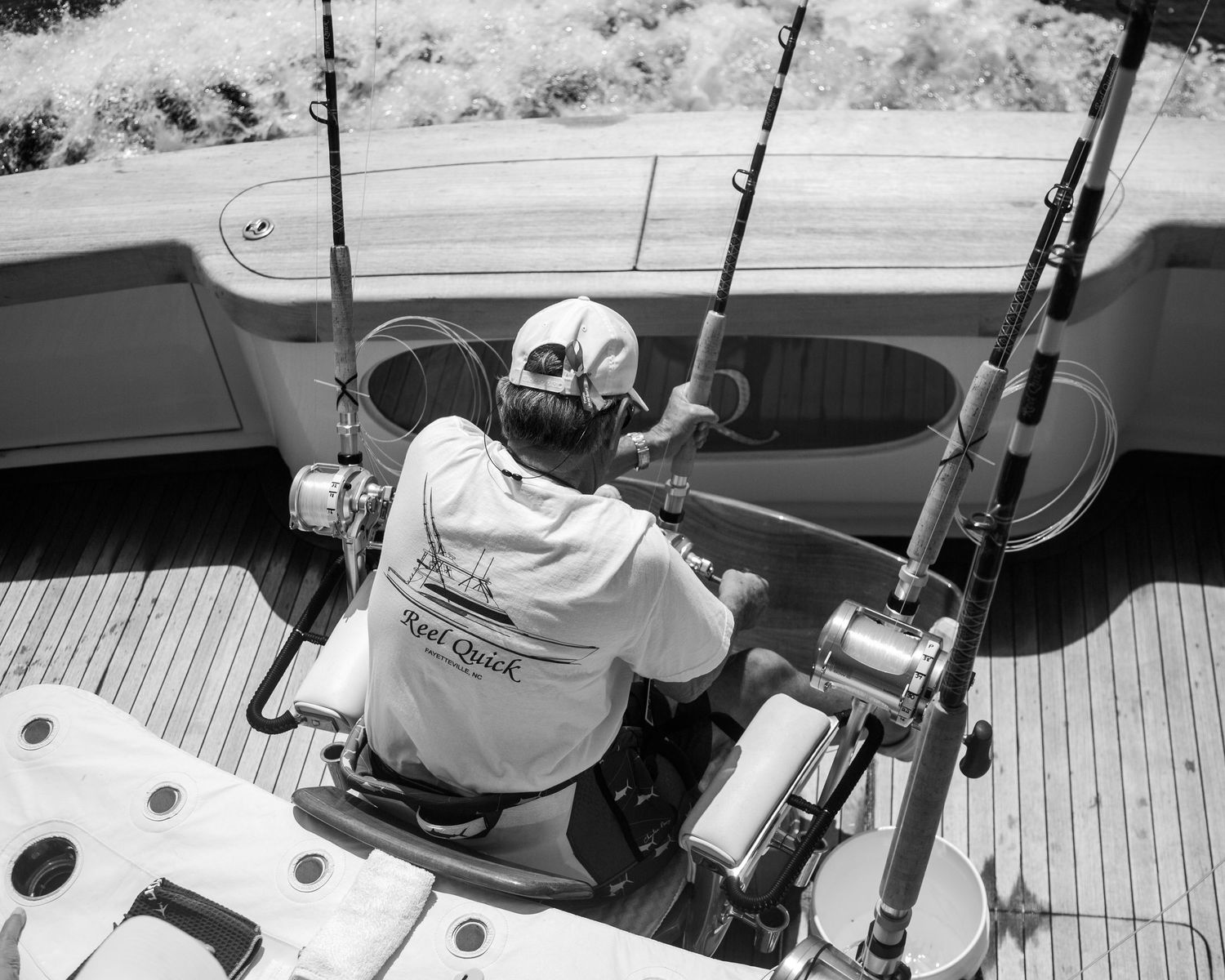 Black and white photo of a fisherman seated on a boat deck surrounded by fishing rods and reels on a sunny day.