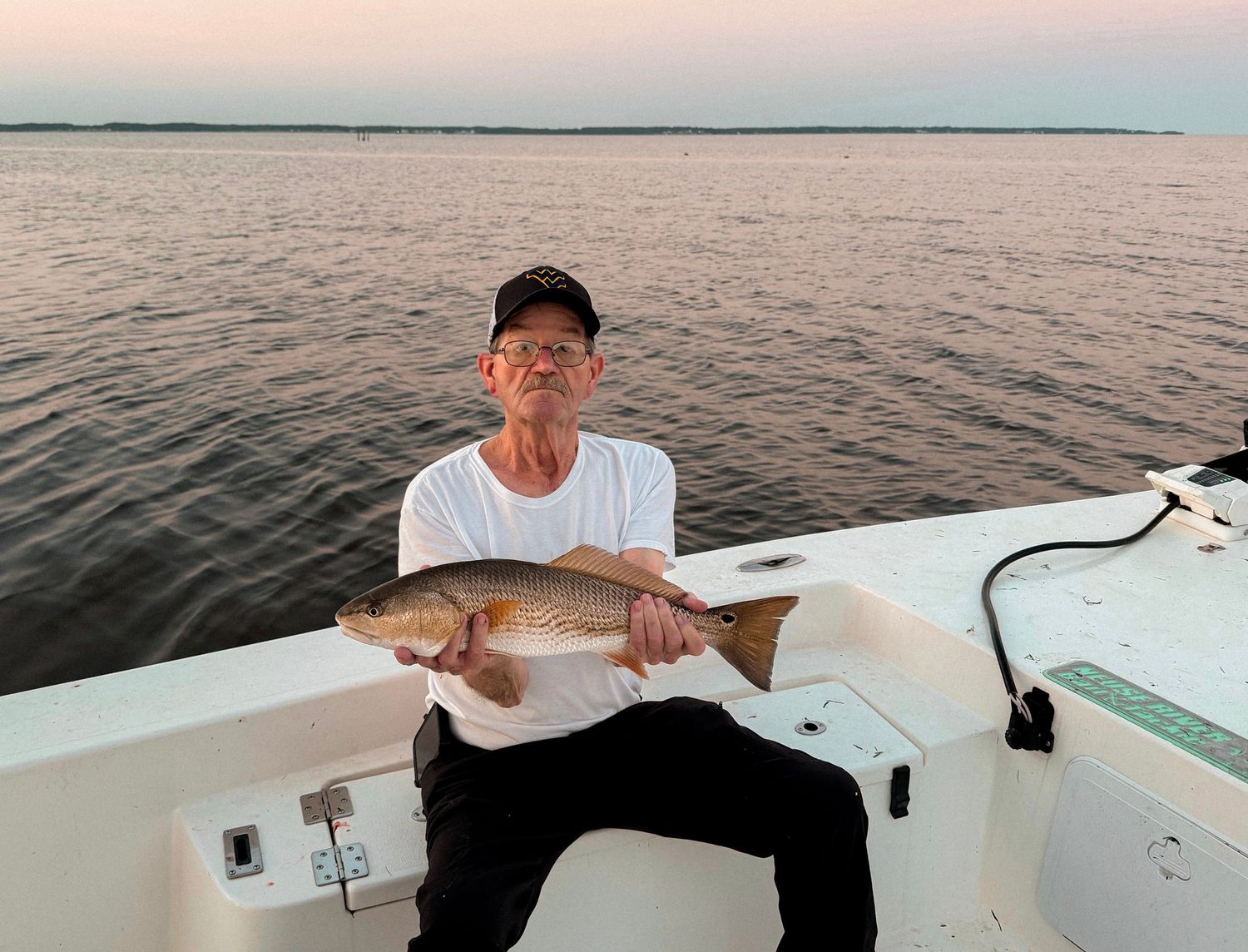 Man holding a large redfish on a boat at sunset over calm coastal waters.
