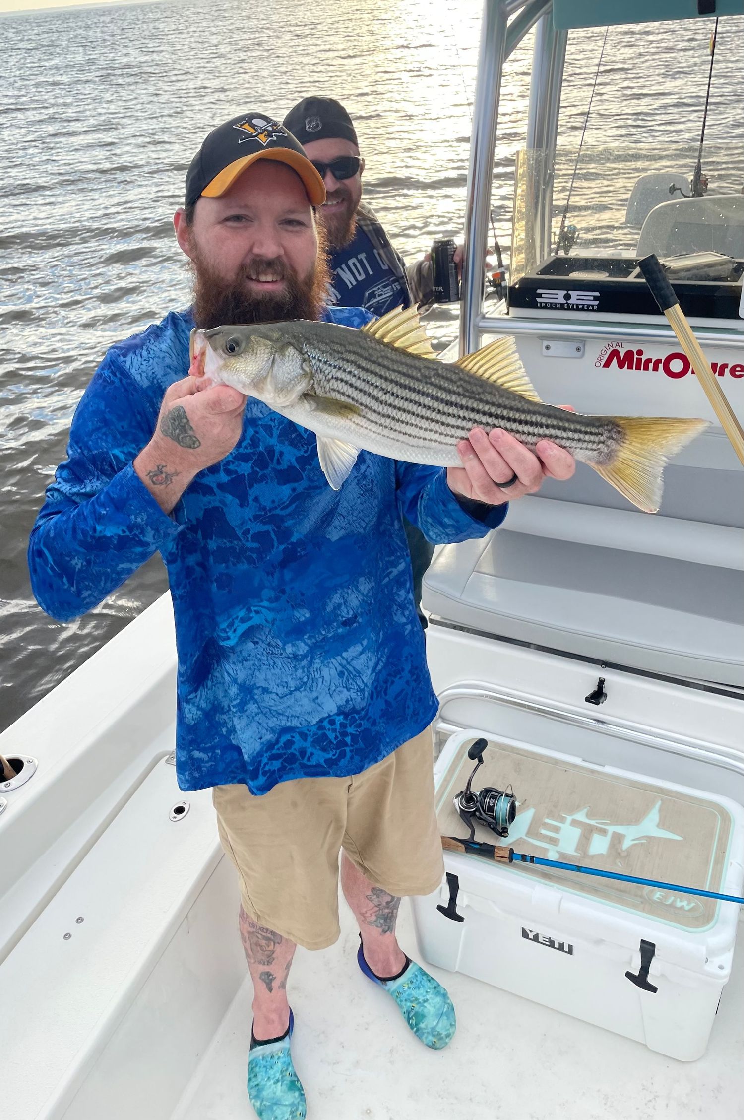 Angler in blue shirt shows off freshly caught striped bass on boat deck during sunset fishing trip.