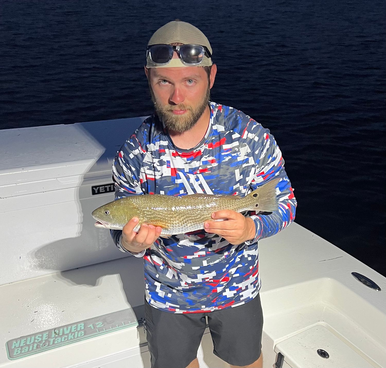 Angler displays freshly caught fish while wearing patriotic camo pattern shirt on boat deck during fishing trip.