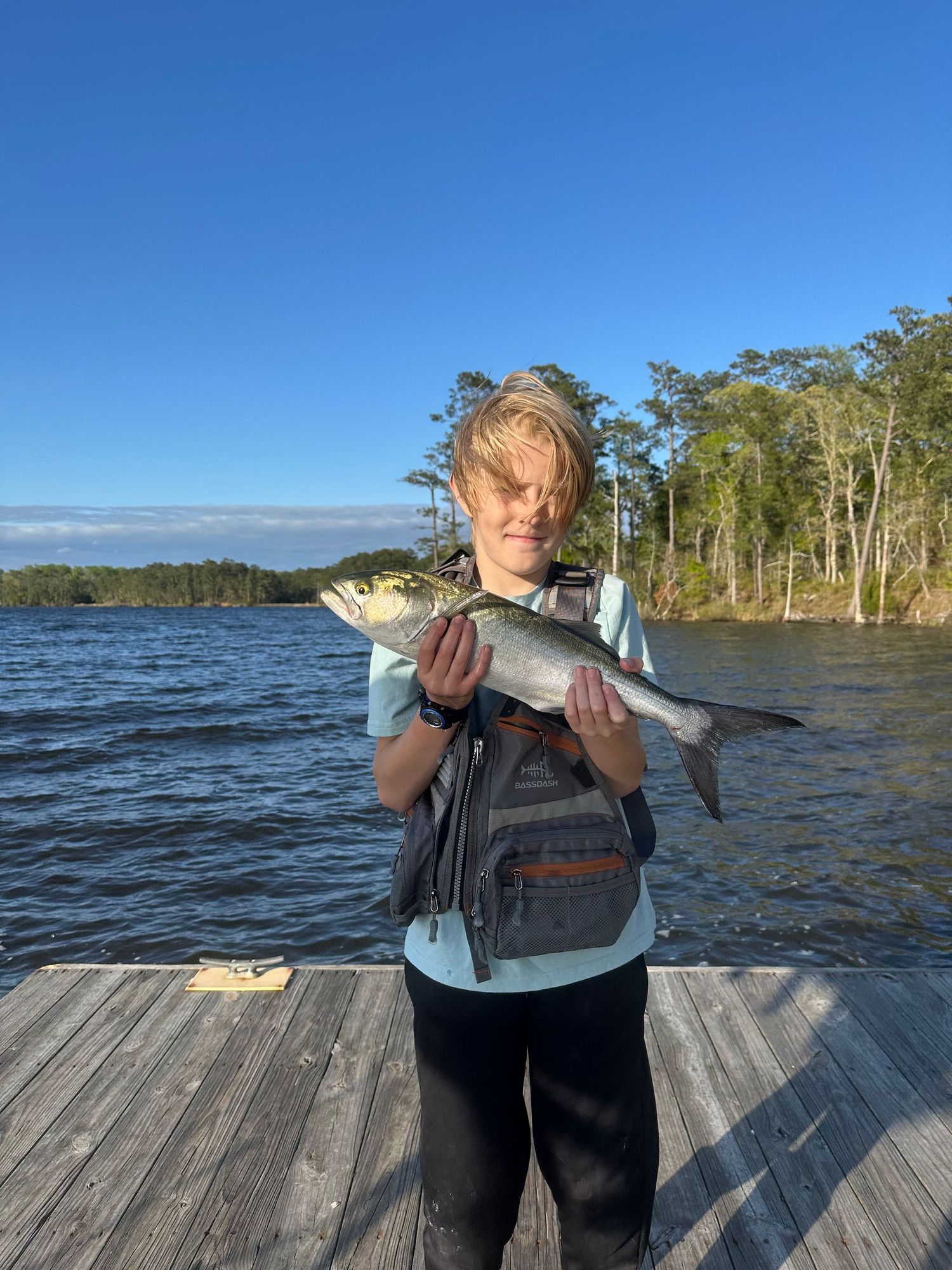 Young angler holding a striped bass catch on wooden dock with waterway and trees in background.