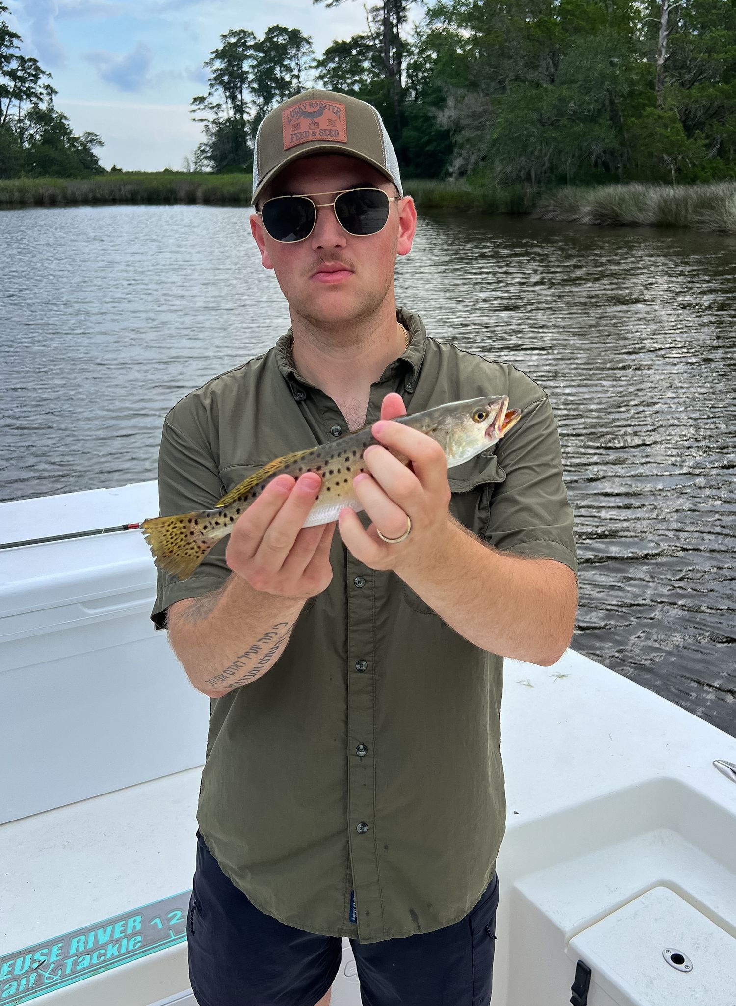 Man in olive shirt and cap holds a spotted trout on a boat near a marshy river.