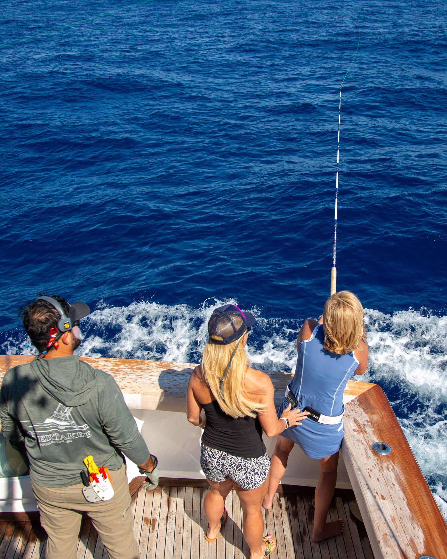 Three people fishing from the stern of a boat on deep blue ocean waters, with one person holding a tall fishing rod.