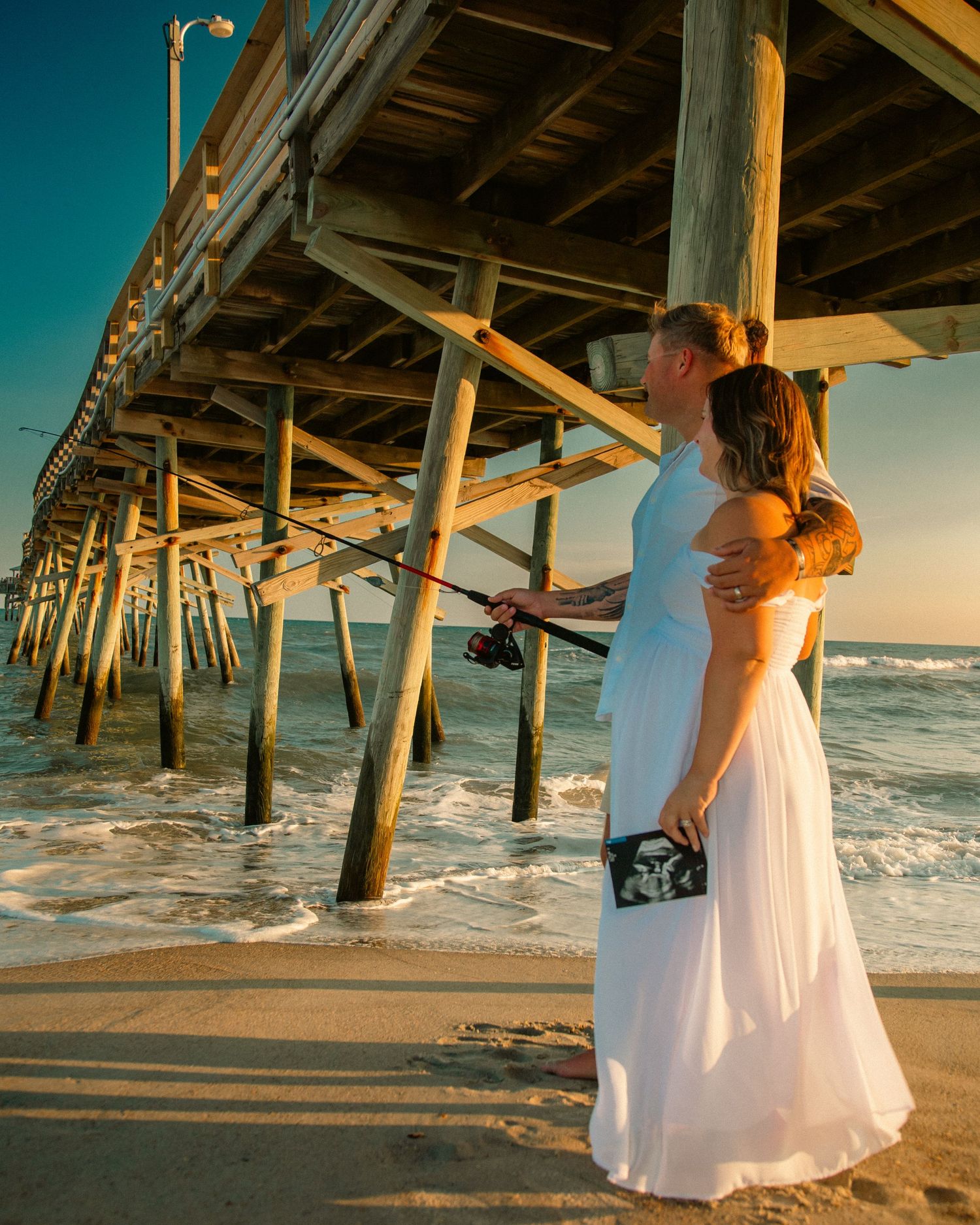 Couple in wedding attire embracing on beach under wooden pier at sunset, groom holding fishing rod.