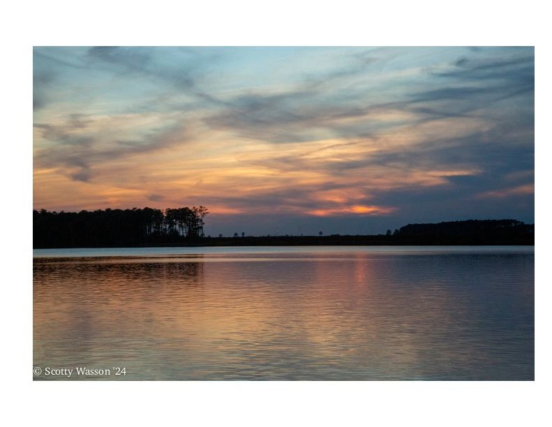 Tranquil sunset over a calm lake with silhouetted trees along the shoreline casting reflections on the water.