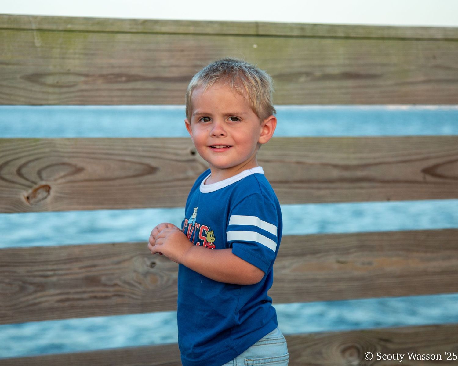A young blond boy in a blue t-shirt smiles while standing in front of a wooden fence near the ocean.
