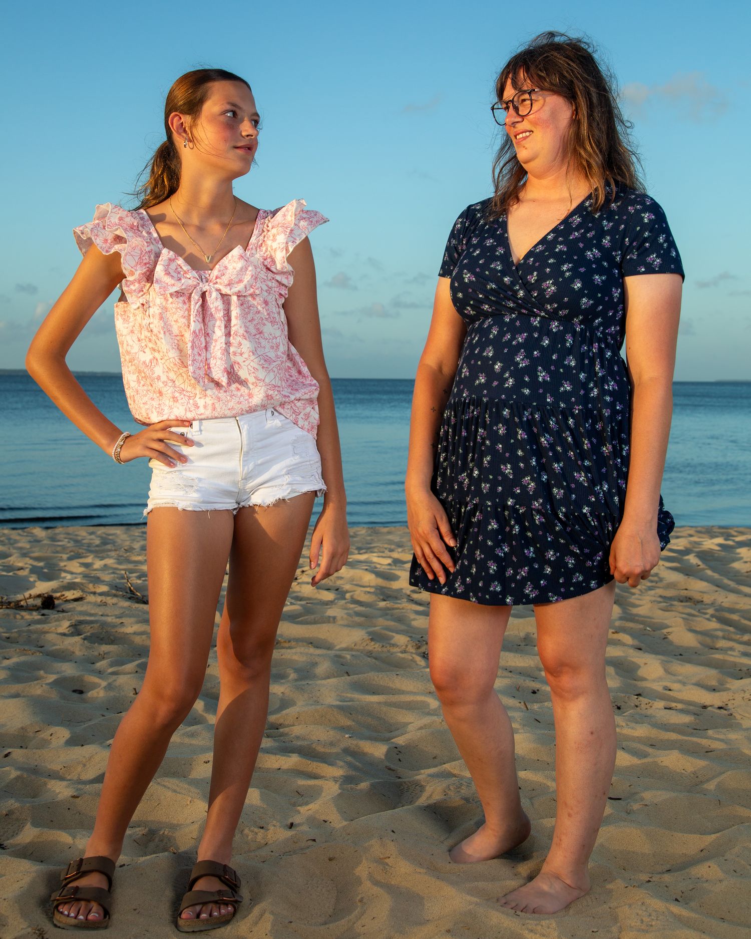 Two women standing on a sandy beach at sunset, one in pink floral top and shorts, other in navy floral dress.