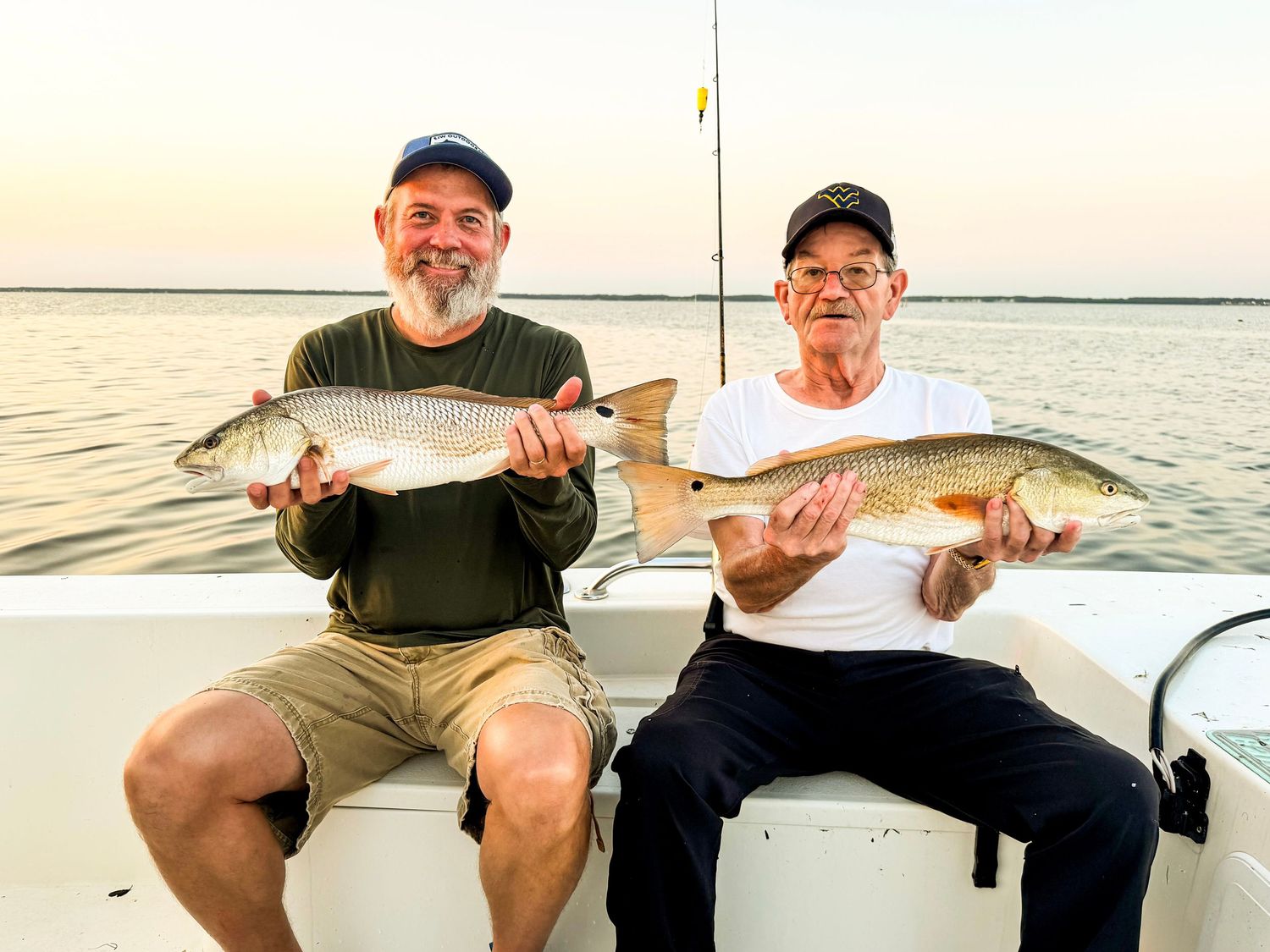 Two men sitting on a boat proudly holding large redfish caught while fishing on calm waters at sunset.