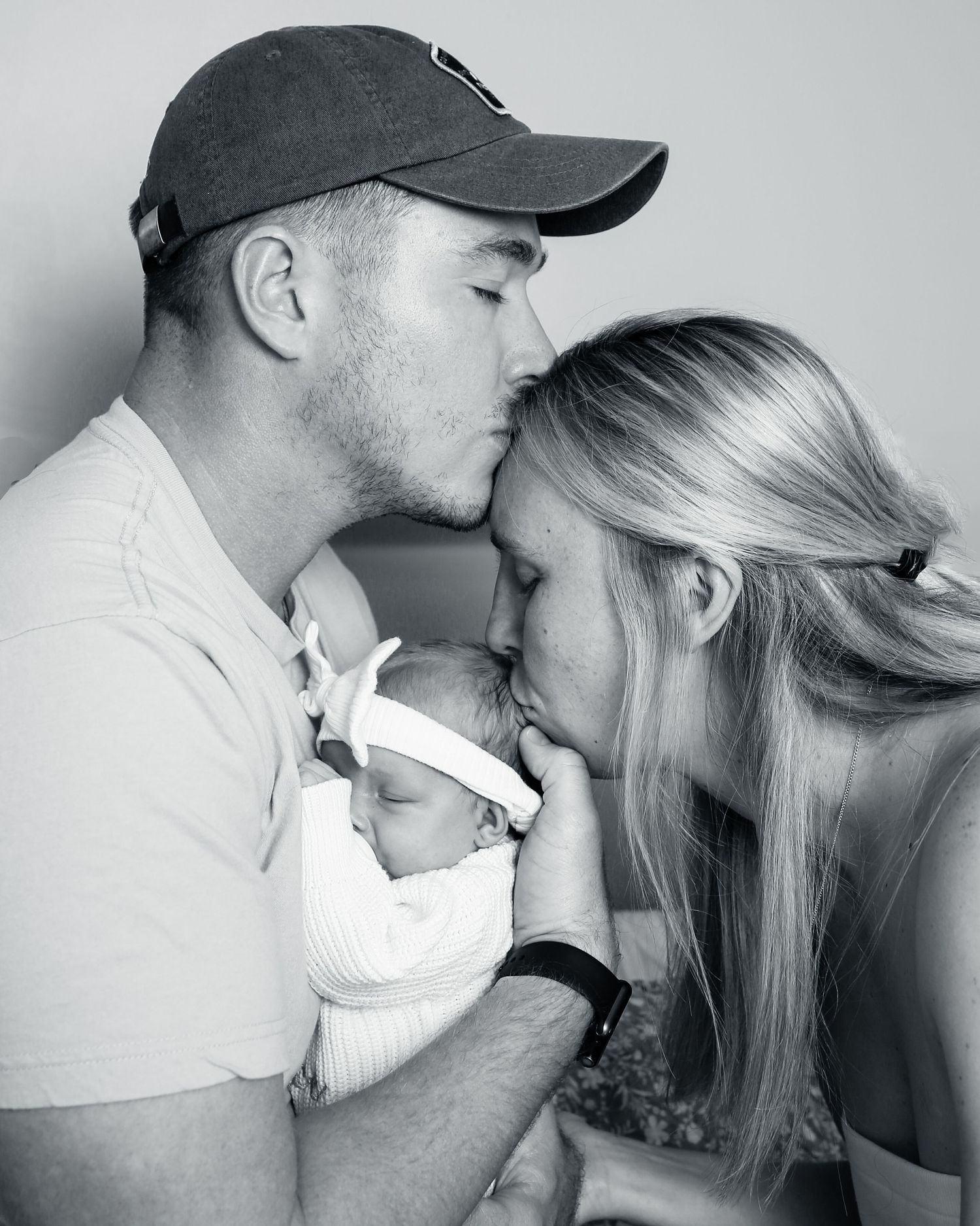 Black and white intimate family portrait showing gentle moment with newborn baby held between two people in tender embrace.