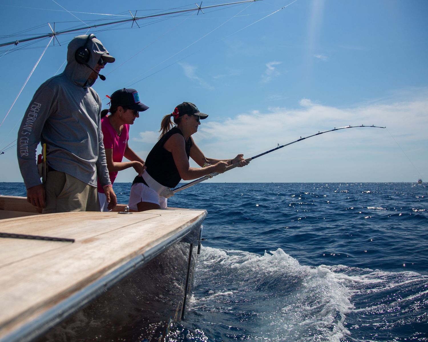 Three people fishing from a boat on a sunny day, with a woman reeling in a catch over the deep blue ocean.