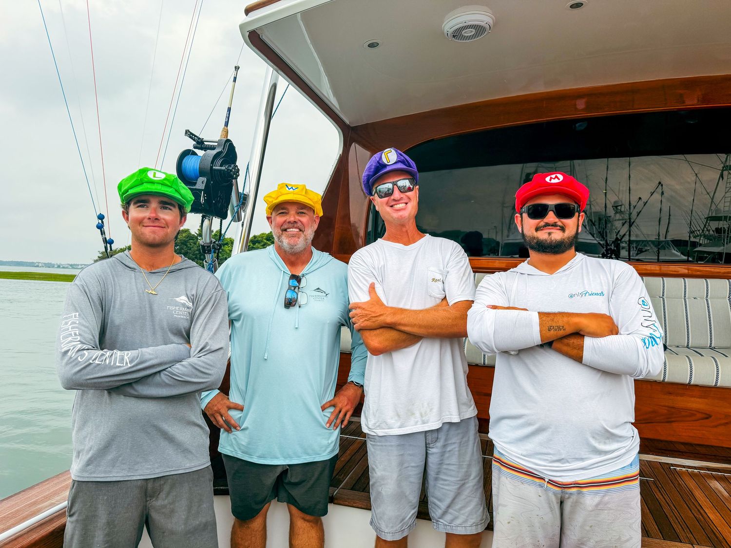 Four men in colorful hats pose together on a boat deck near calm waters on a cloudy day.