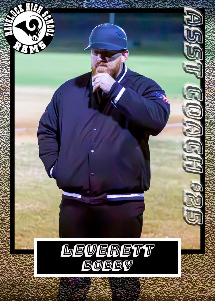 Baseball coach in navy uniform and blue cap stands thoughtfully on field during evening game.