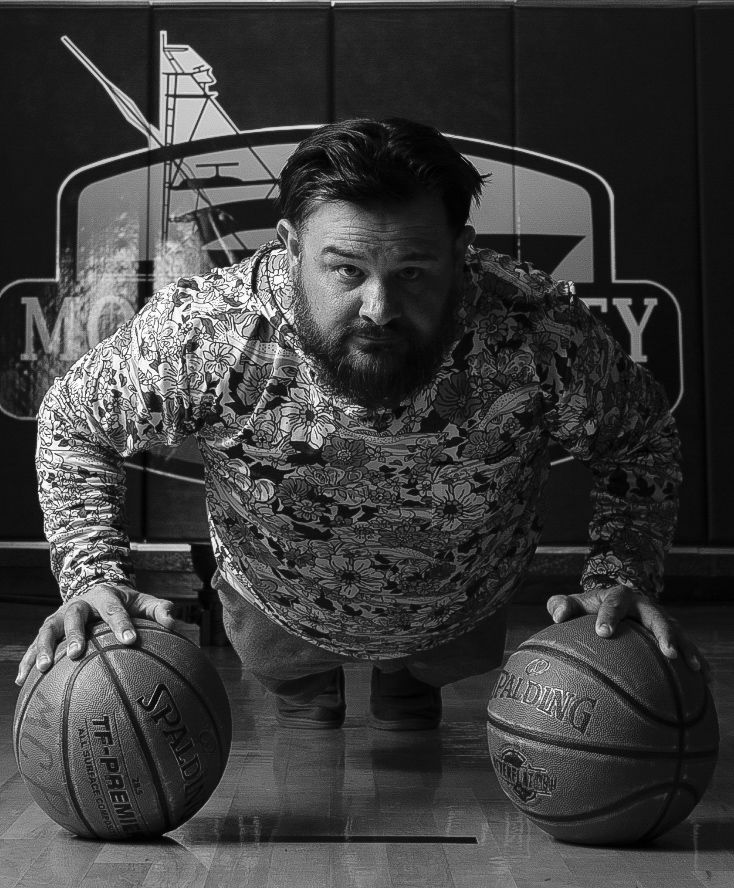 Black and white photo of a muscular athlete posing with two basketballs against a gym backdrop.
