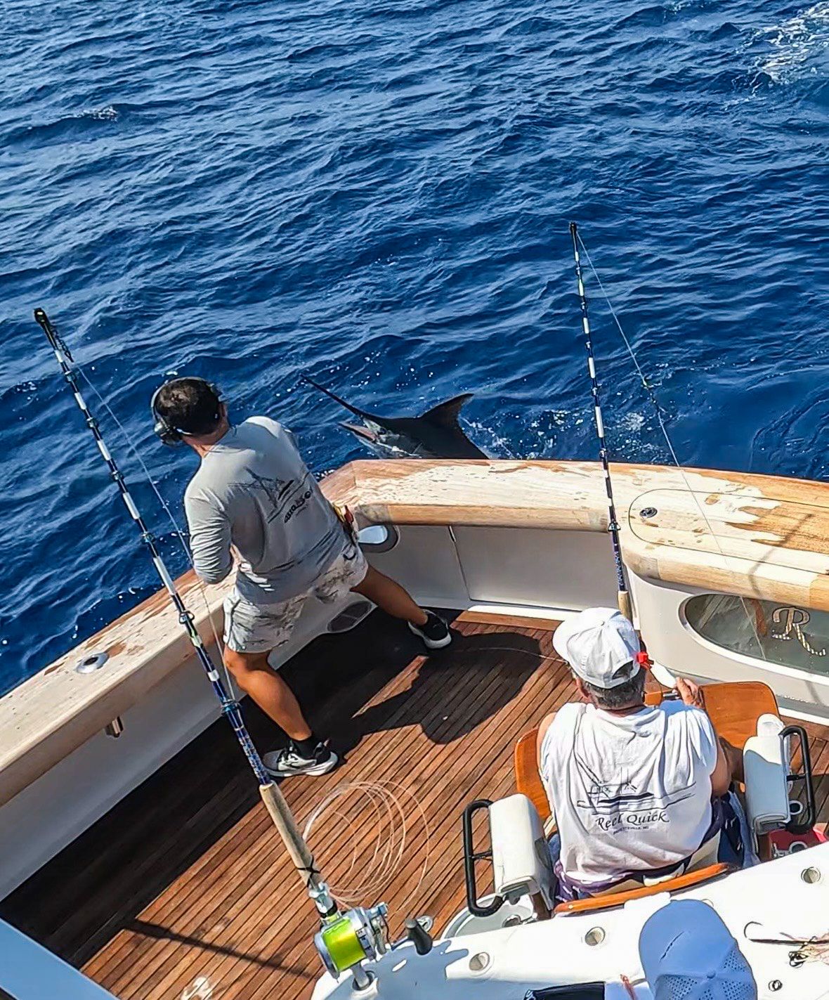 Two anglers on a deep sea fishing boat battle a marlin in blue ocean waters with multiple fishing rods rigged.