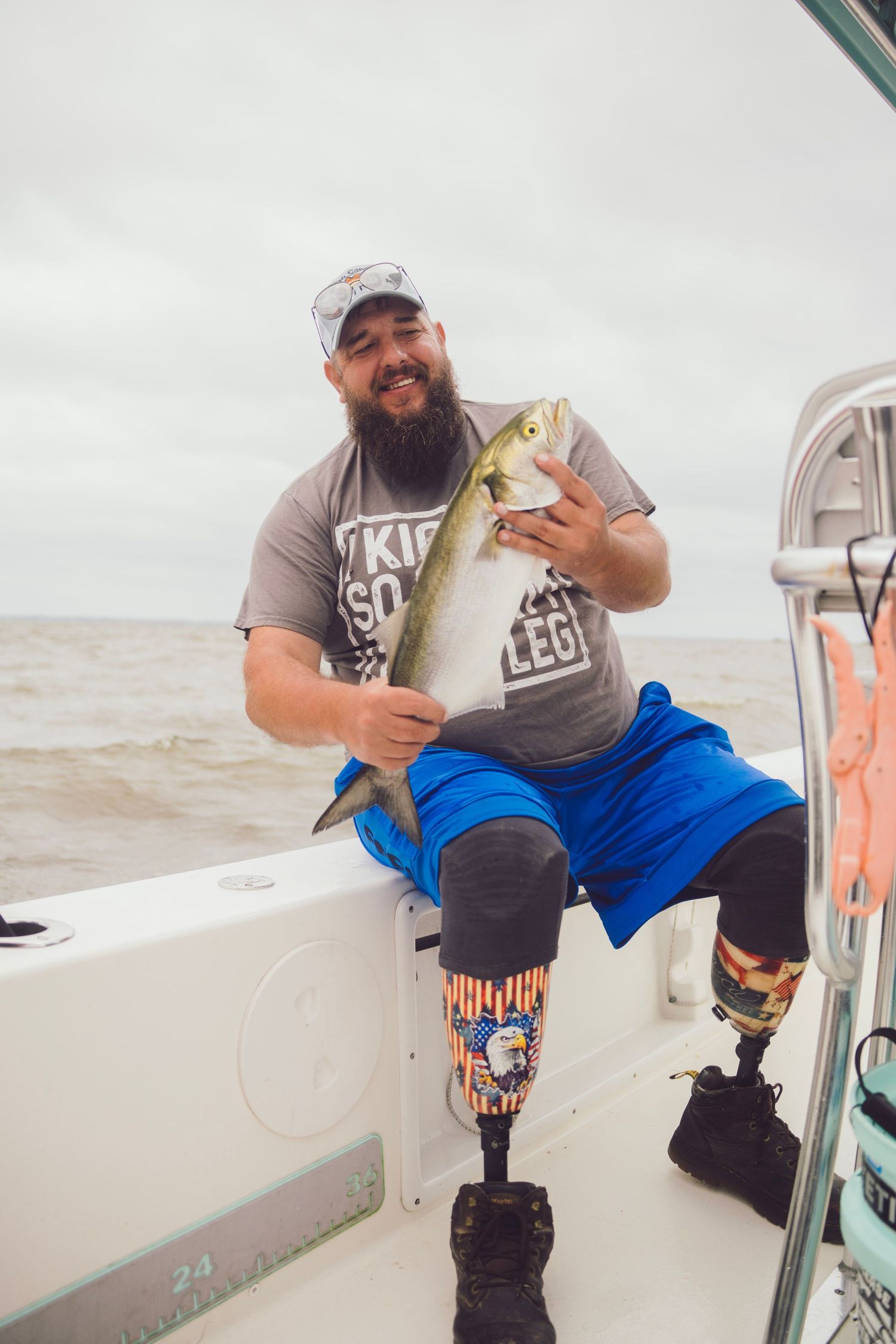 Smiling bearded man with prosthetic legs holds a fish on a boat, wearing blue shorts and a gray t-shirt.