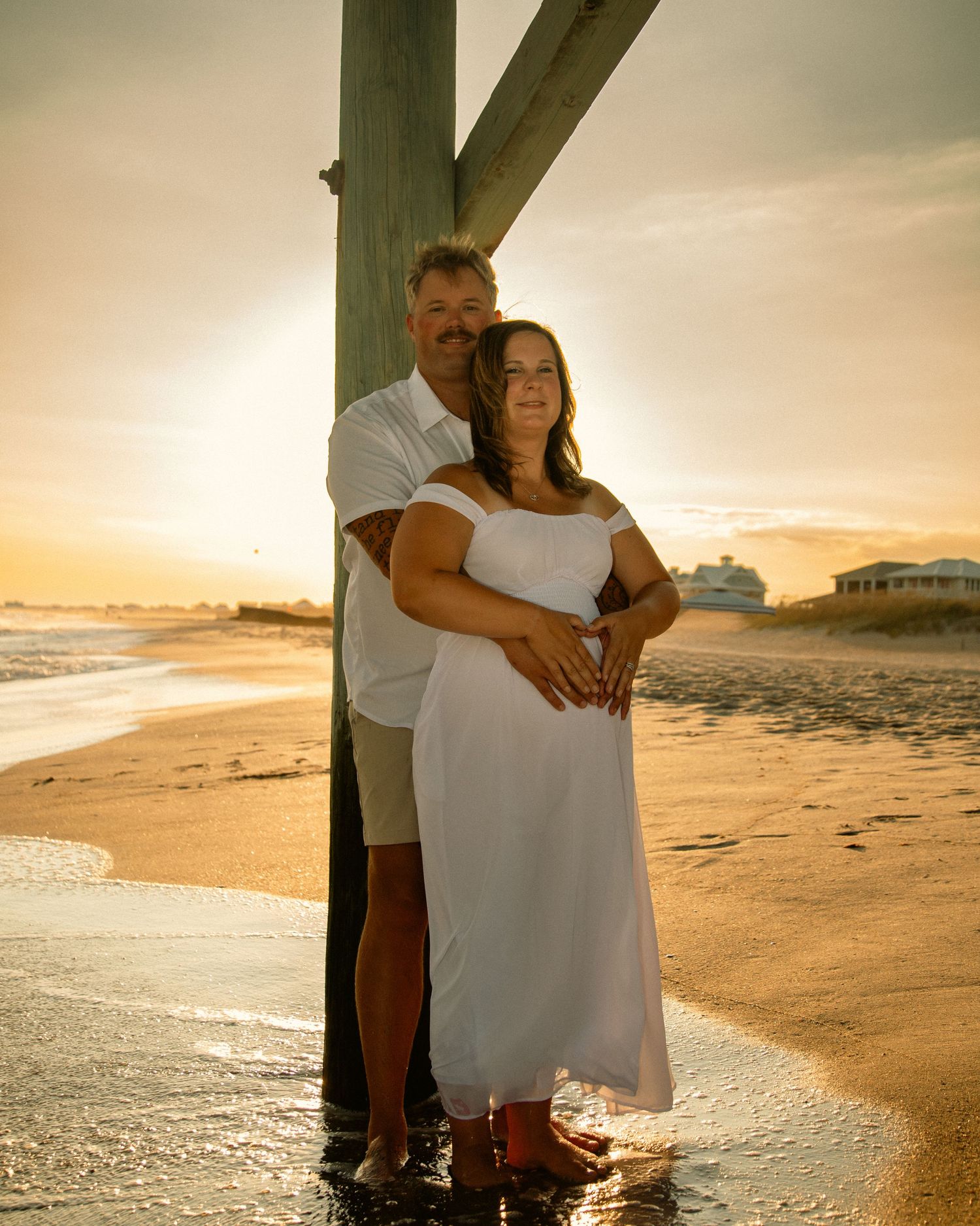 Couple embracing at beach sunset near wooden post, woman in white dress, golden hour ocean backdrop.