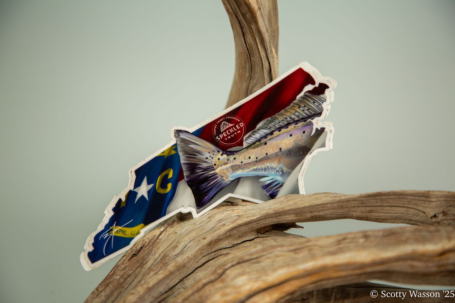 A colorful fish-shaped sticker featuring the North Carolina flag design, displayed on driftwood against a gray background.