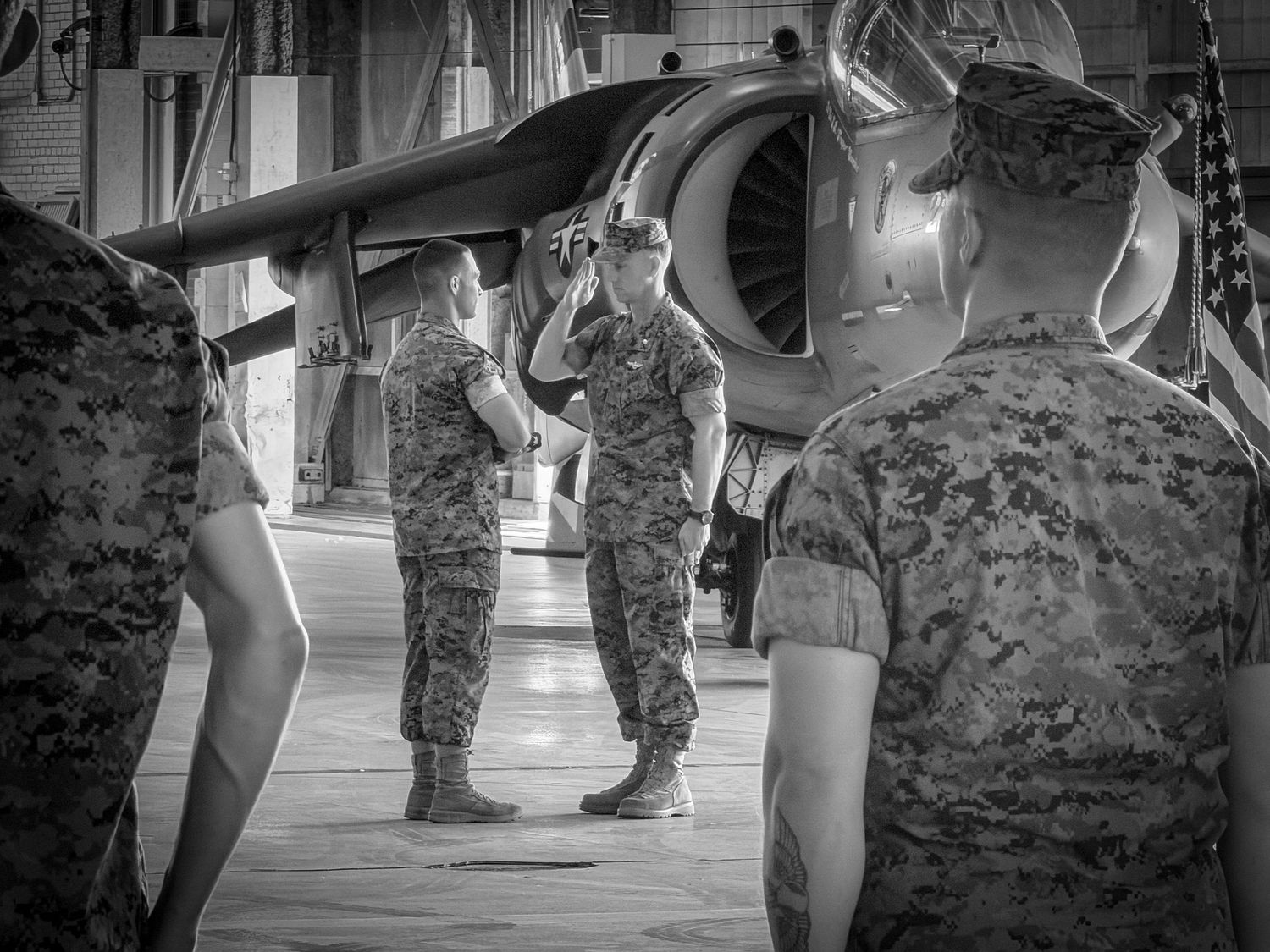 Marines in camouflage uniforms saluting near a military aircraft inside a hangar, black and white photo.