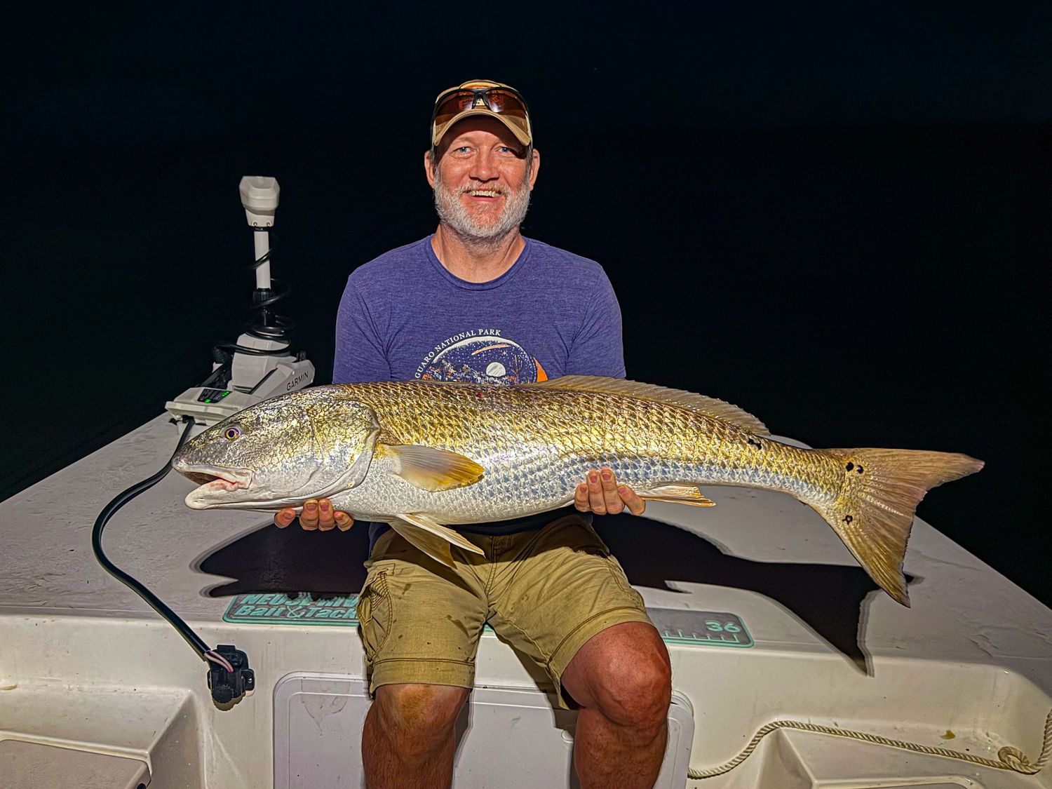 Man proudly holds a large snook fish on a boat at night, showcasing a successful nighttime fishing catch.
