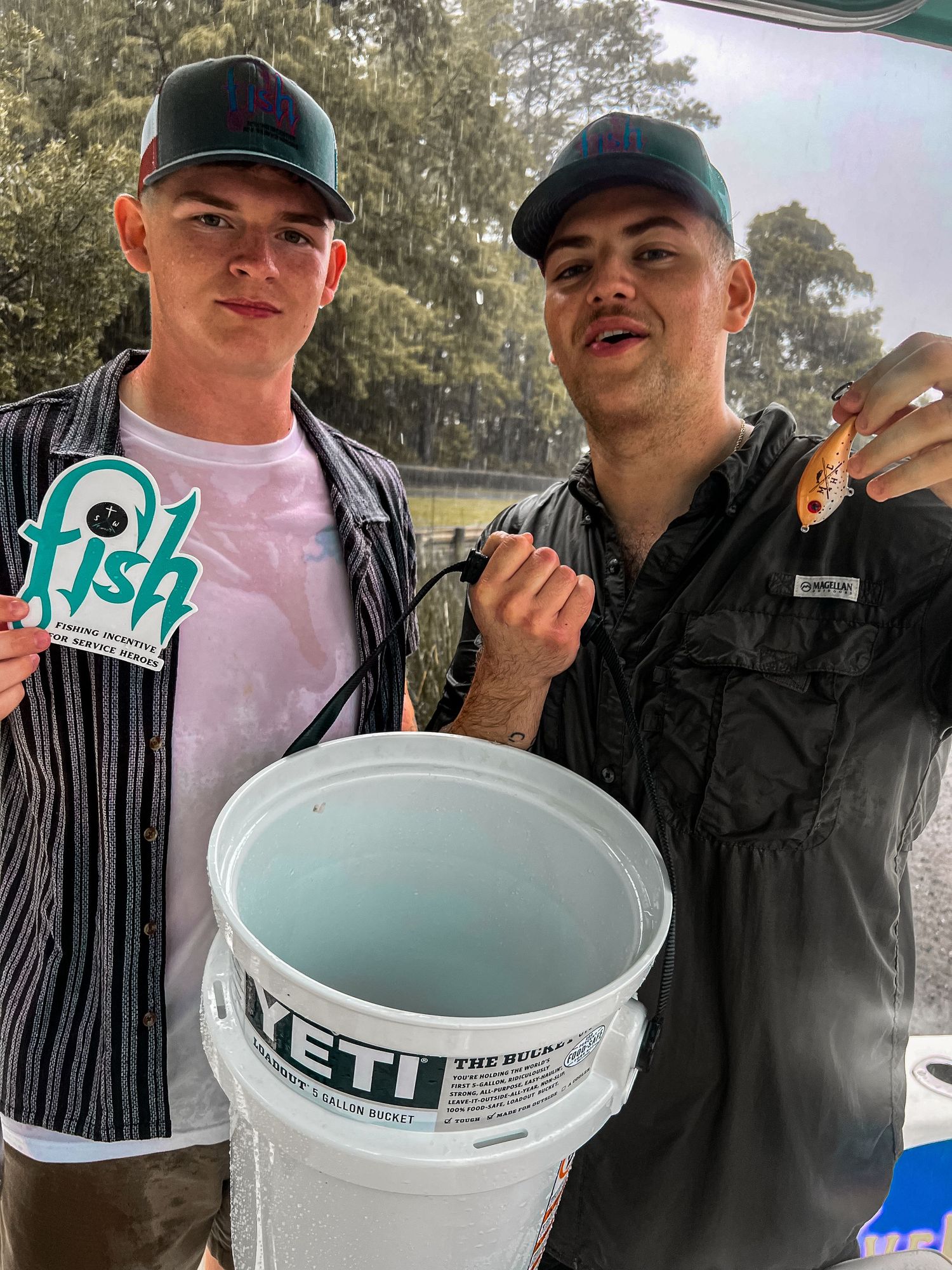 Two young men posing outdoors with a Yeti bucket, one holding a fish lure and wearing a fish logo shirt.