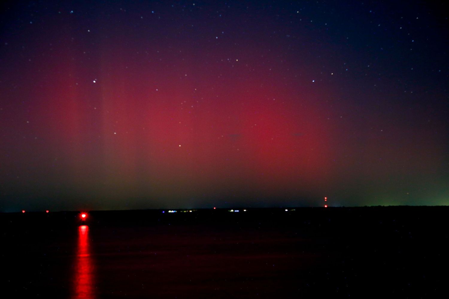 Stunning red and green aurora borealis northern lights display over calm water at night with stars visible.