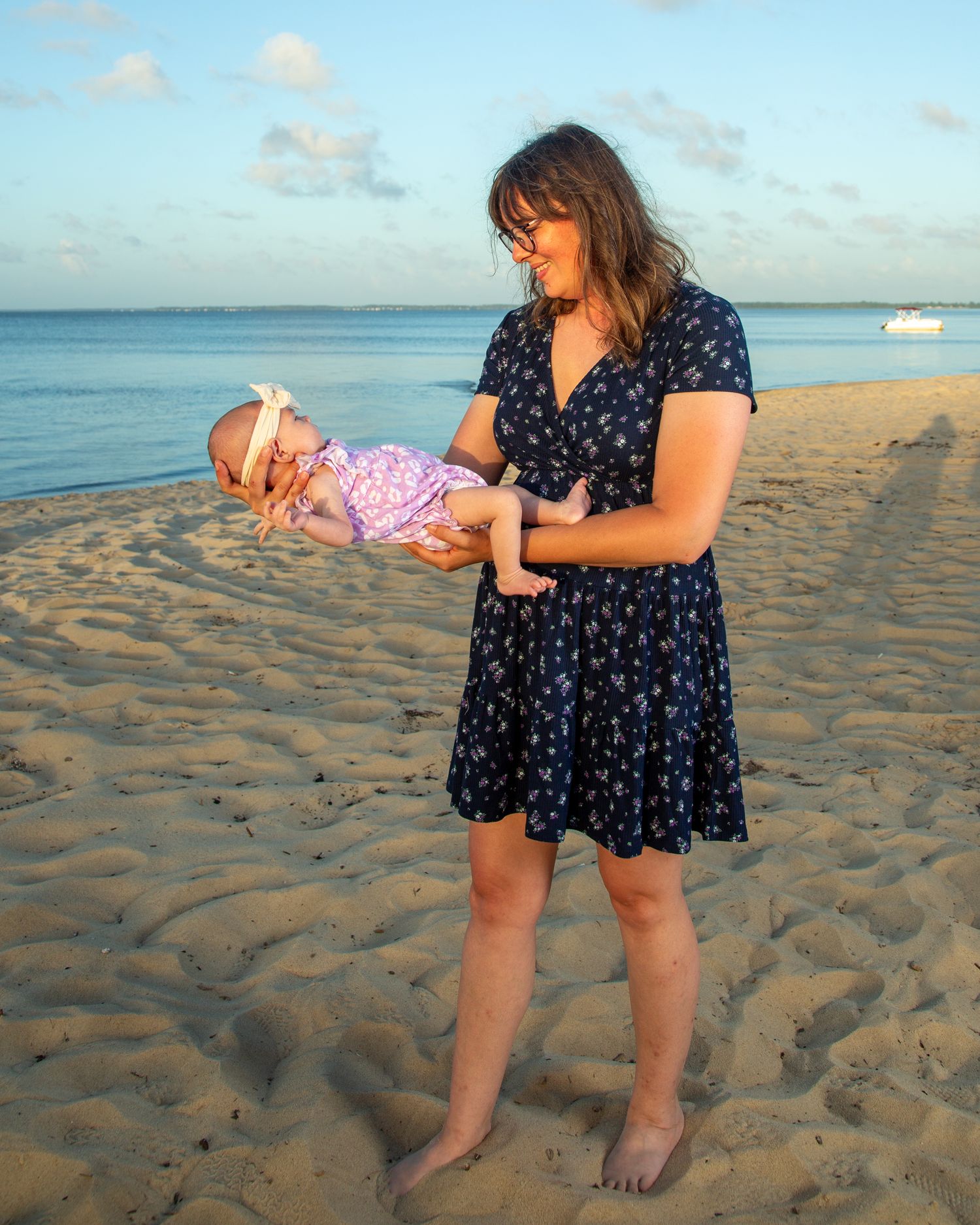 A woman in a navy floral dress holds a baby on a sandy beach at golden hour with calm ocean waters behind her.
