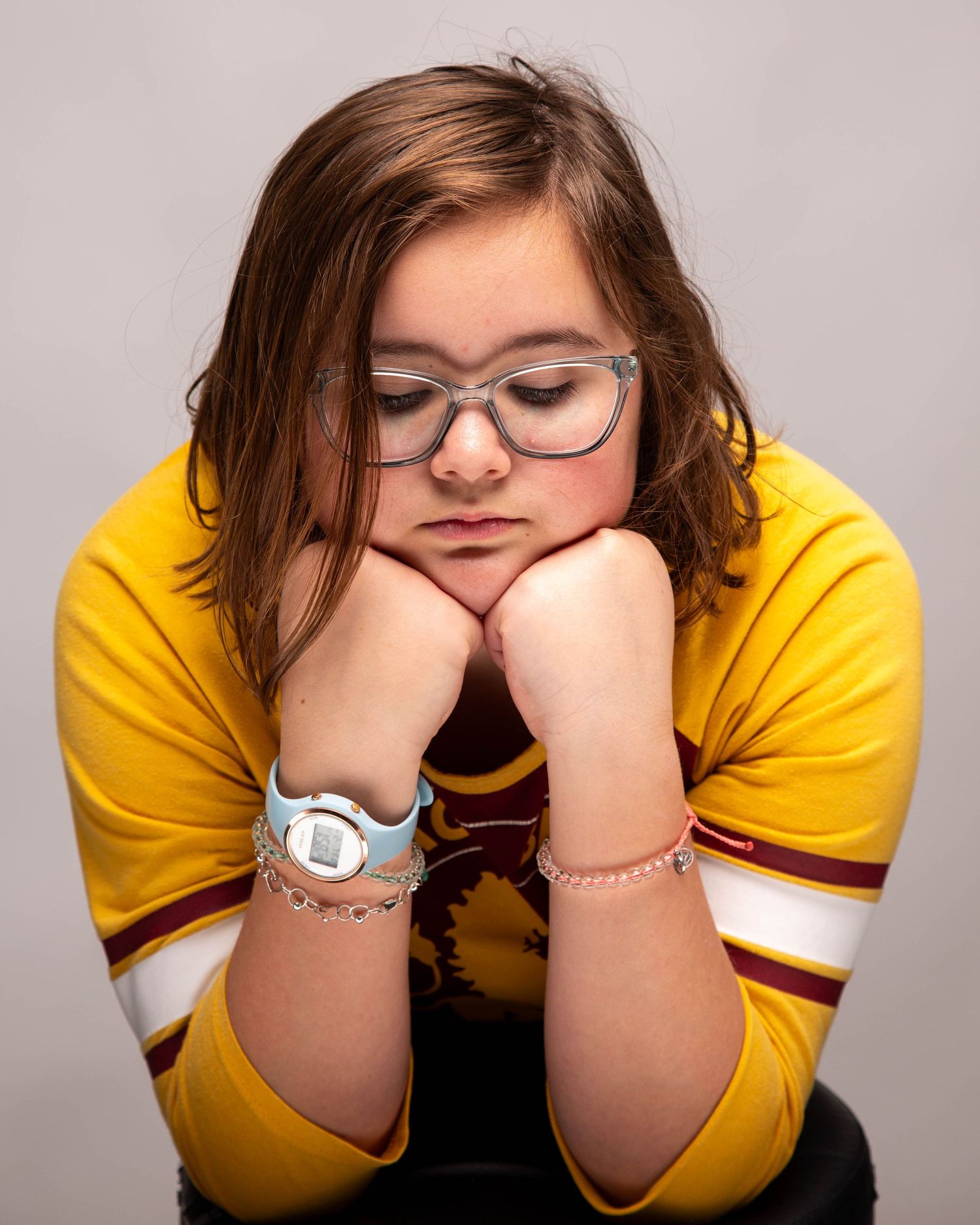 Young girl with glasses and yellow sweater rests chin on hands, looking thoughtful in a studio portrait.