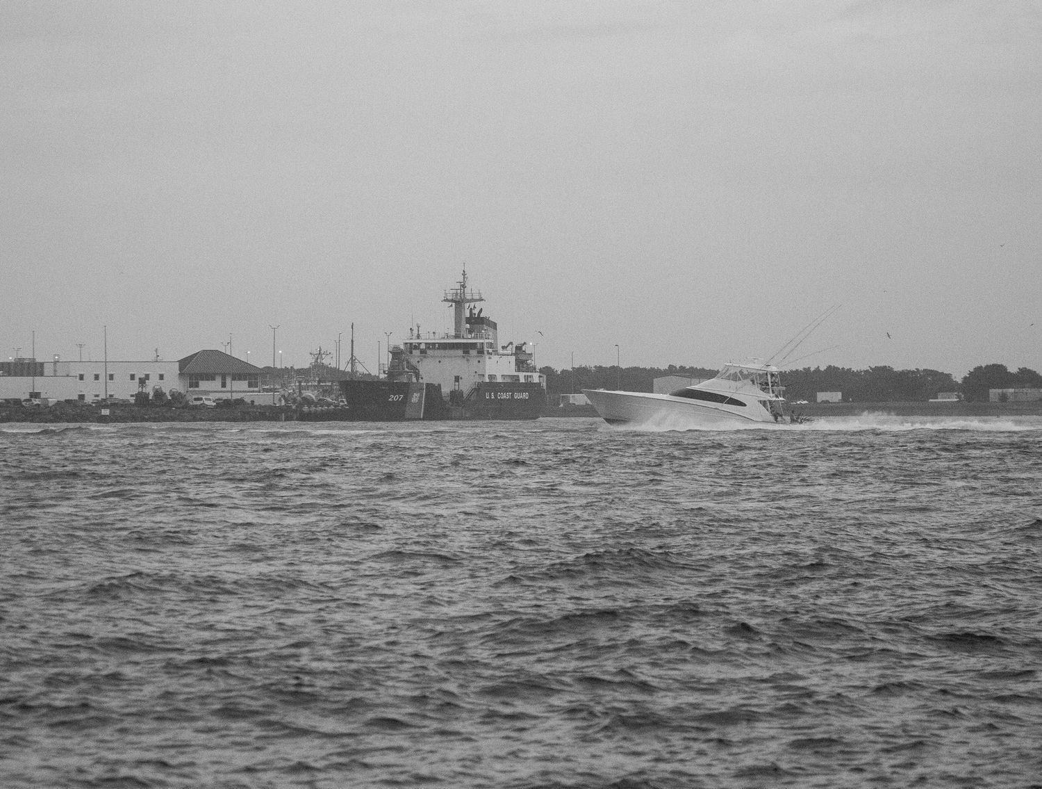 Historic lighthouse and coastal structures viewed from the water in black and white.