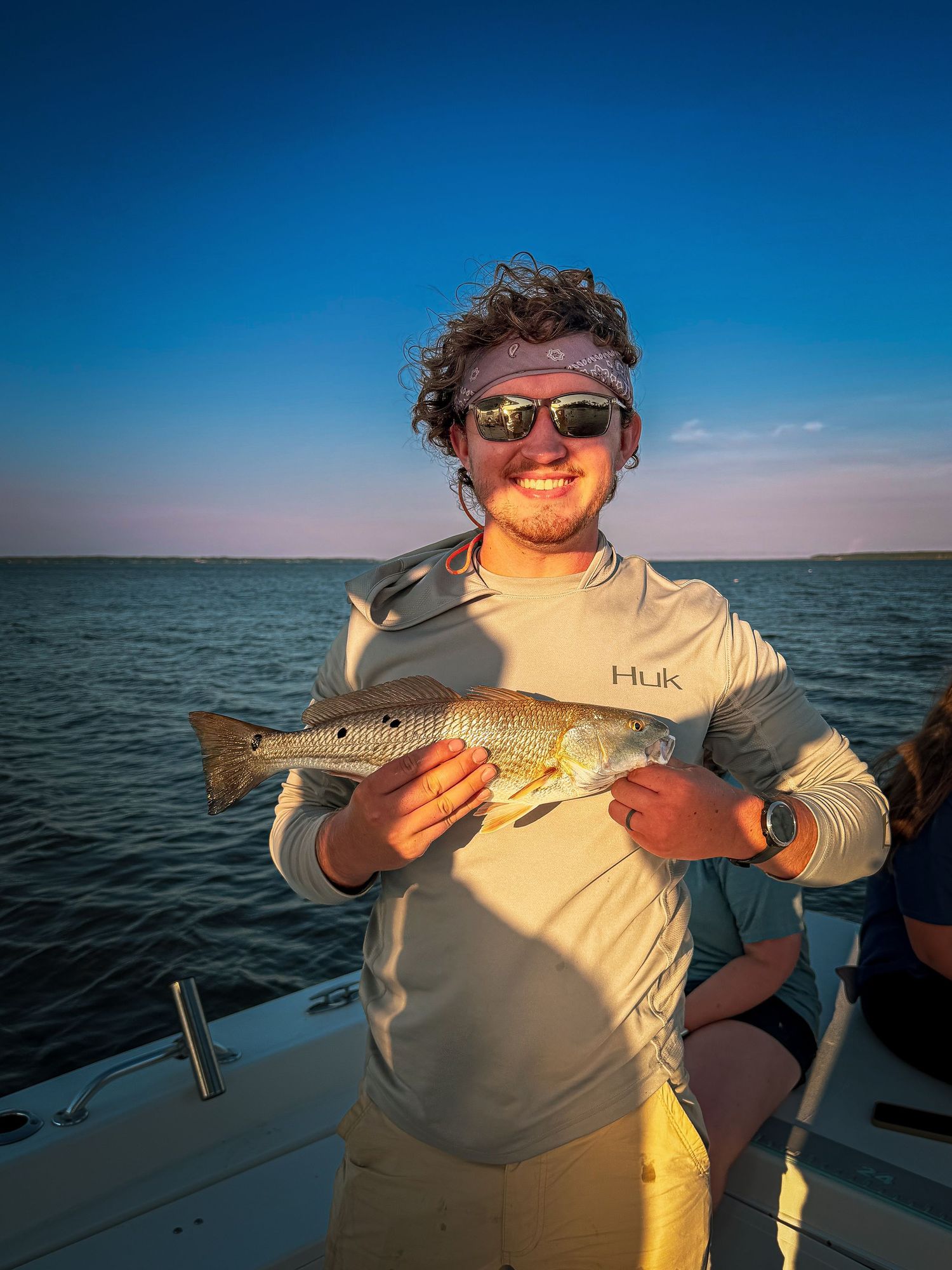 Smiling young man in Huk shirt and sunglasses holding a redfish on a boat at sunset over calm water.