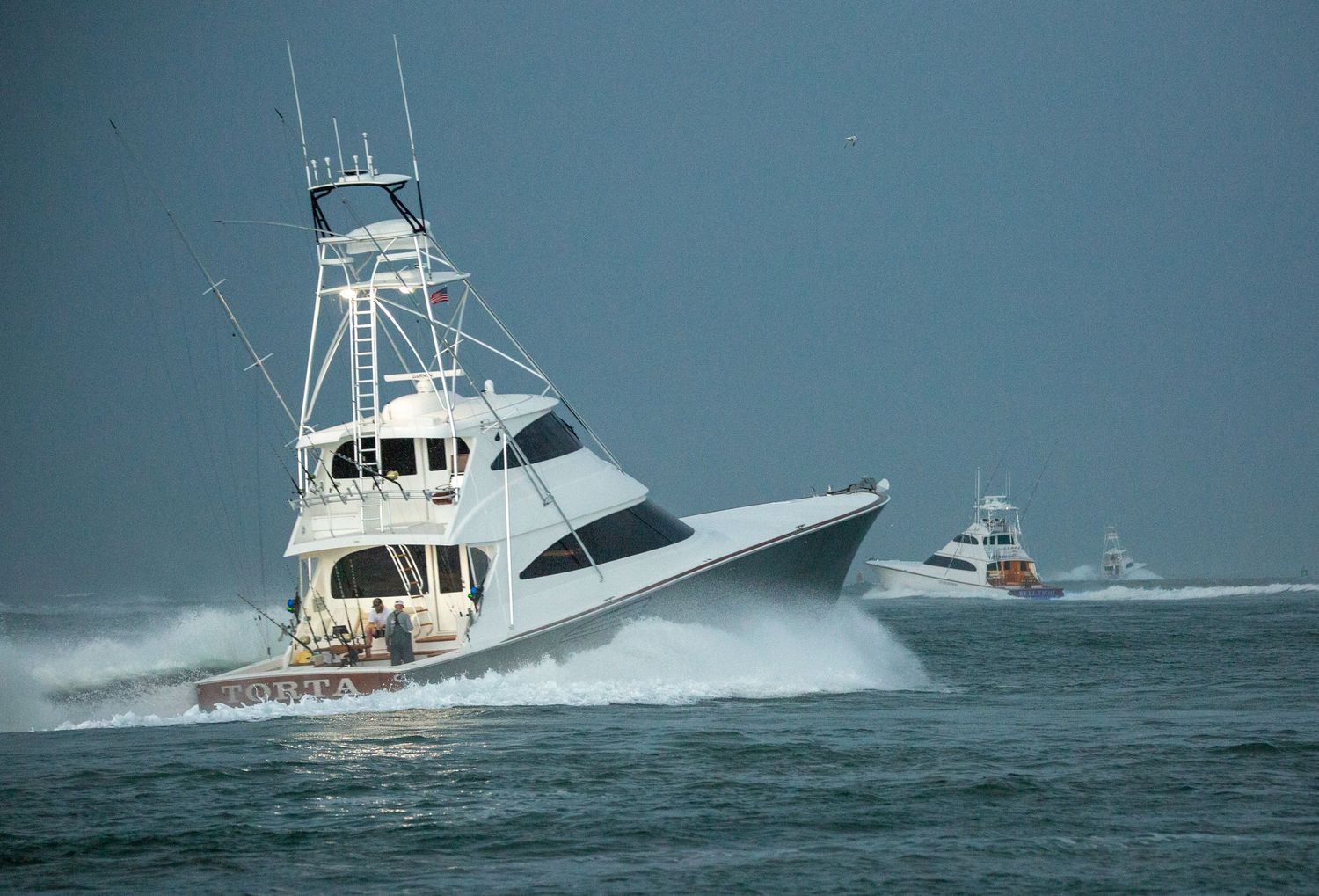 White sportfishing vessel navigating through rough seas with another boat visible in background.