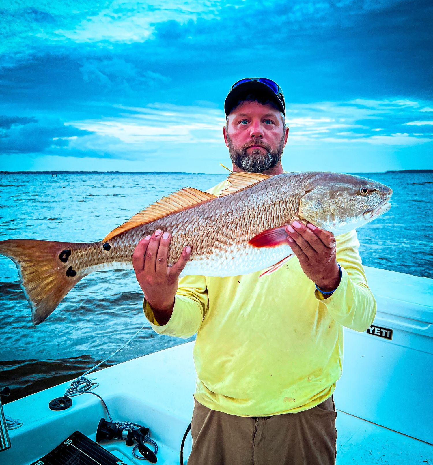 Angler in high-visibility yellow shirt proudly displays large redfish catch on boat against vibrant blue ocean backdrop.
