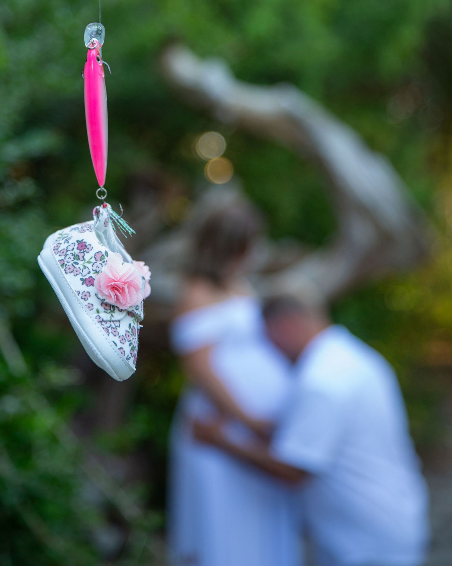 A pink fishing lure holds a floral baby shoe, with a couple embracing softly in the blurred background outdoors.