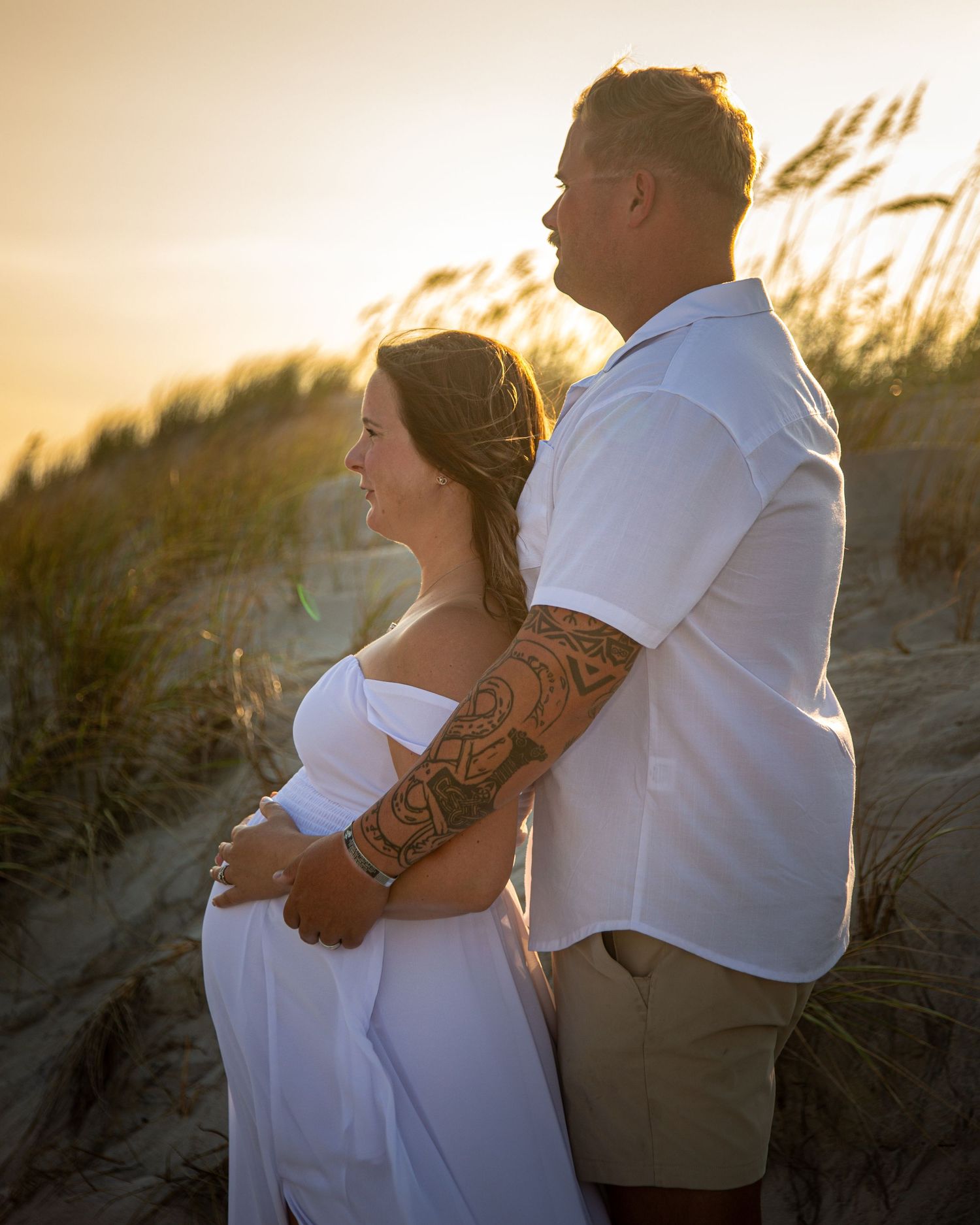 Couple embracing at golden hour on beach dunes, woman in white off-shoulder dress, man with tattoed arm, sunset backdrop.