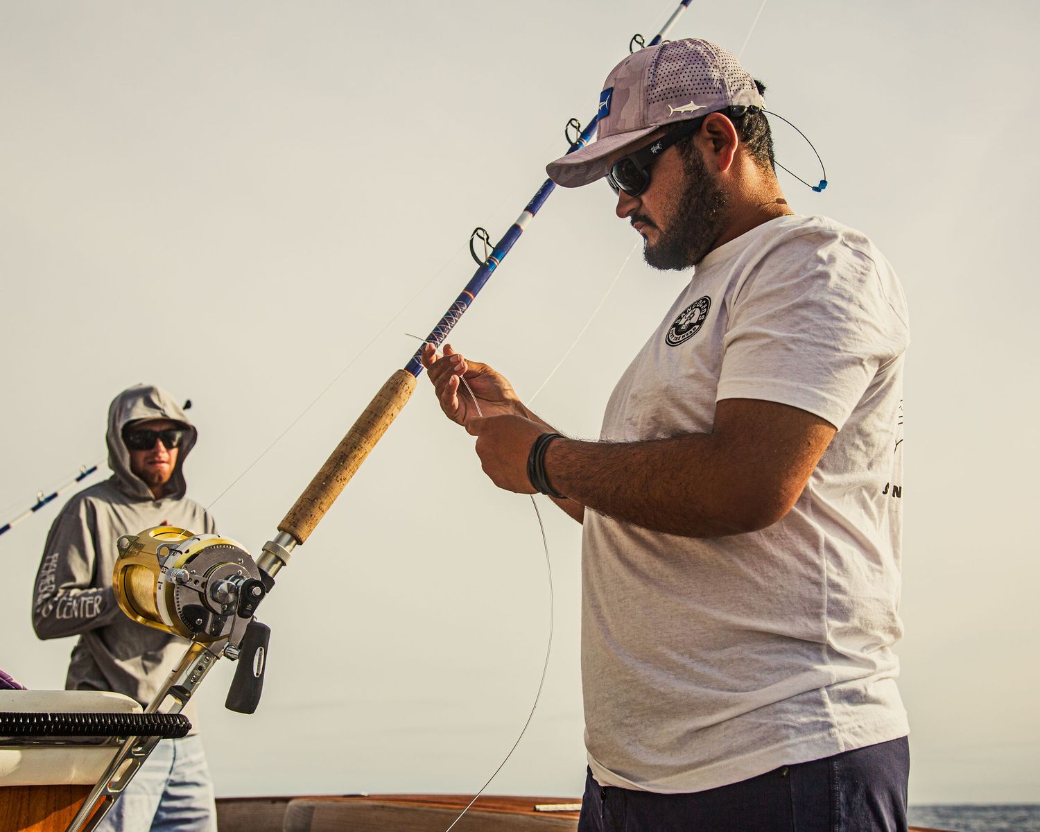 Two men on a boat fishing with large rods in open sea, one examining his fishing line closely.