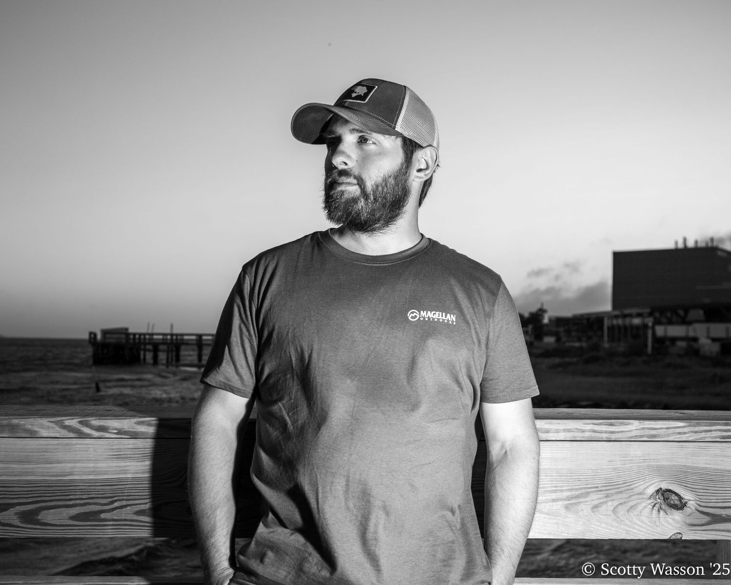 Black and white portrait of bearded man in cap and t-shirt standing outdoors near a pier at dusk.