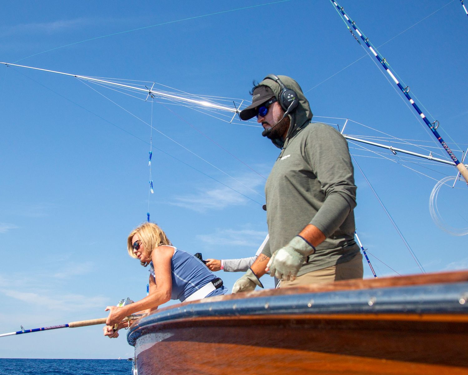 Two anglers fishing from a classic wooden boat on calm blue ocean waters under a sunny sky.