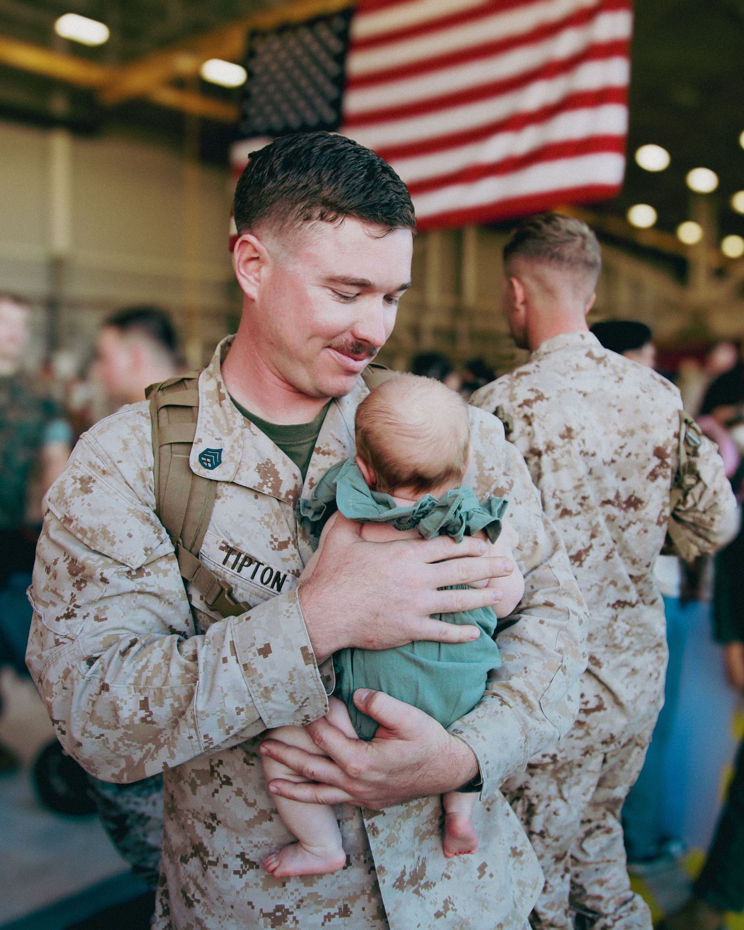 Service member in military uniform holds and comforts a small cat in an indoor setting with American flag visible.