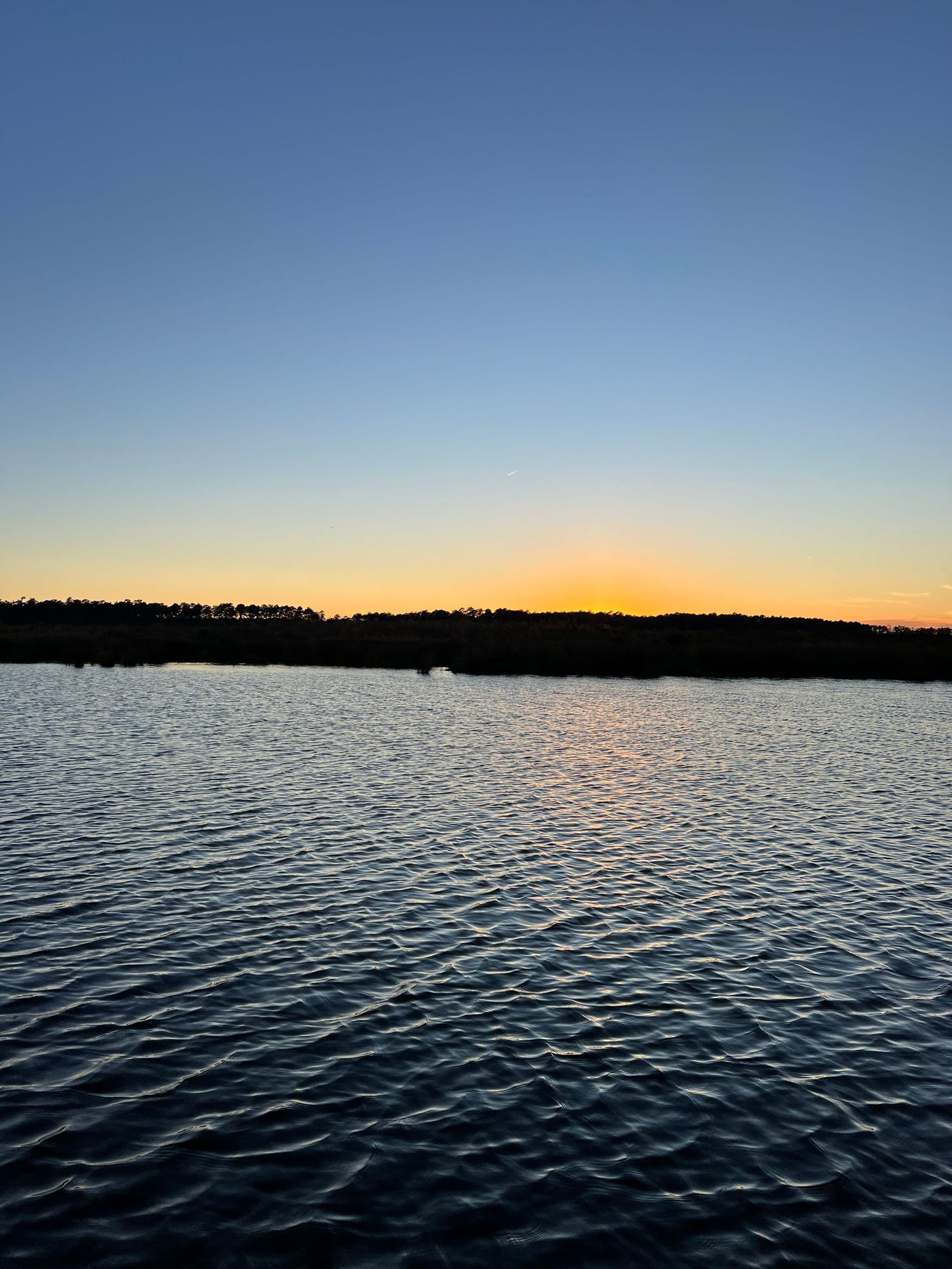 Rippling water surface at sunset with dark tree line silhouette against an orange and blue sky.