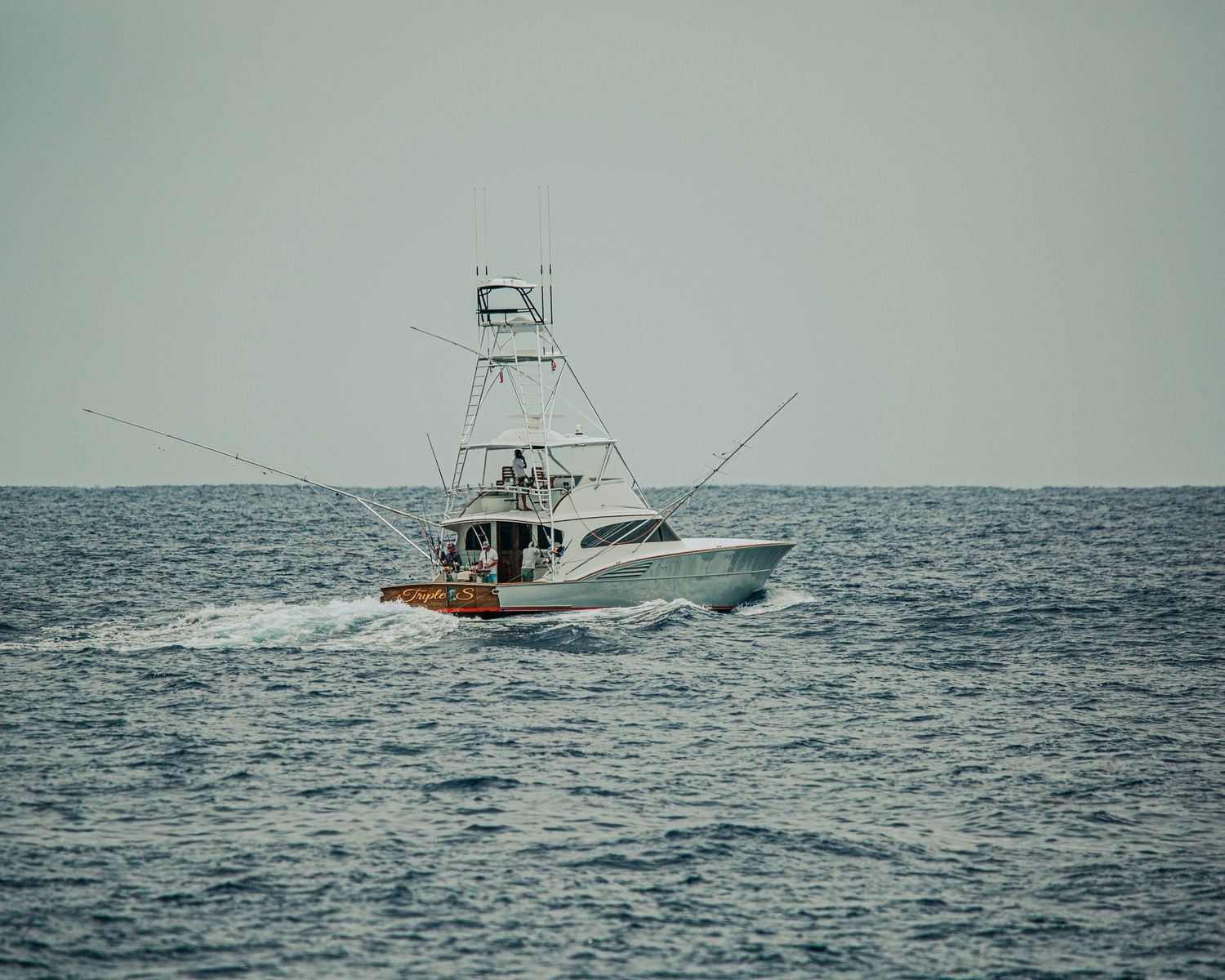 A white sport fishing boat with tall outriggers cruises through blue ocean waters under an overcast sky.