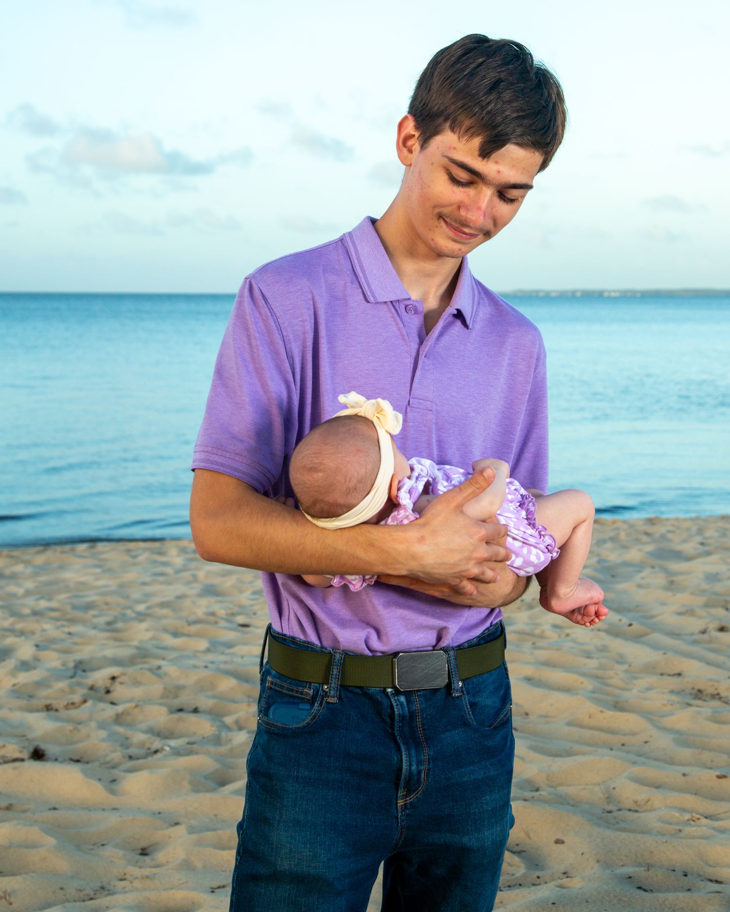 Young man in purple polo shirt tenderly holds a newborn baby on a sandy beach at sunset.