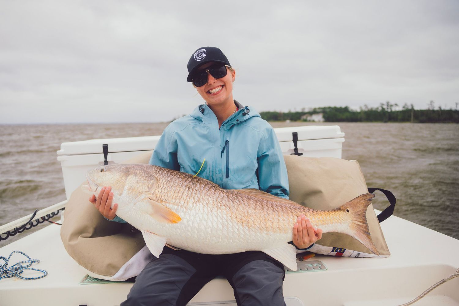 Smiling woman in blue jacket holding a large redfish on a boat on a cloudy day.