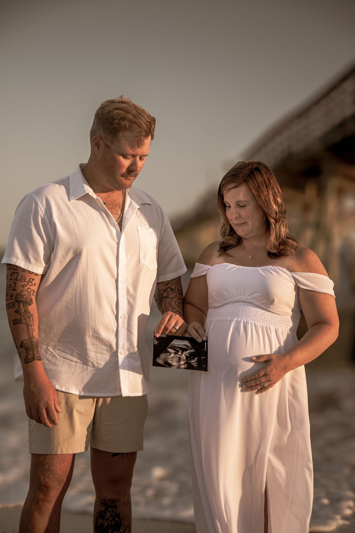 Expecting couple in white outfits holding an ultrasound photo on a beach at sunset near a pier.
