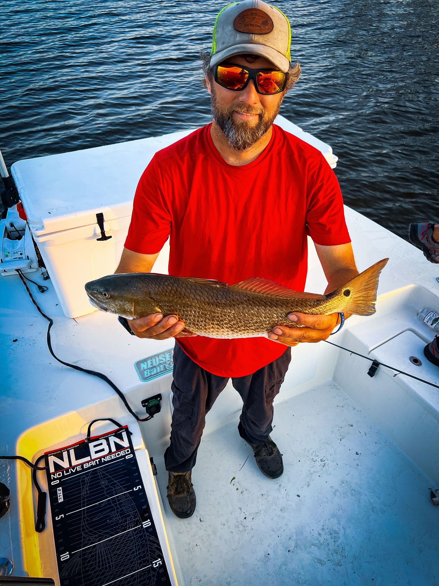 Angler in red shirt displays large redfish catch on boat deck with fishing equipment visible.