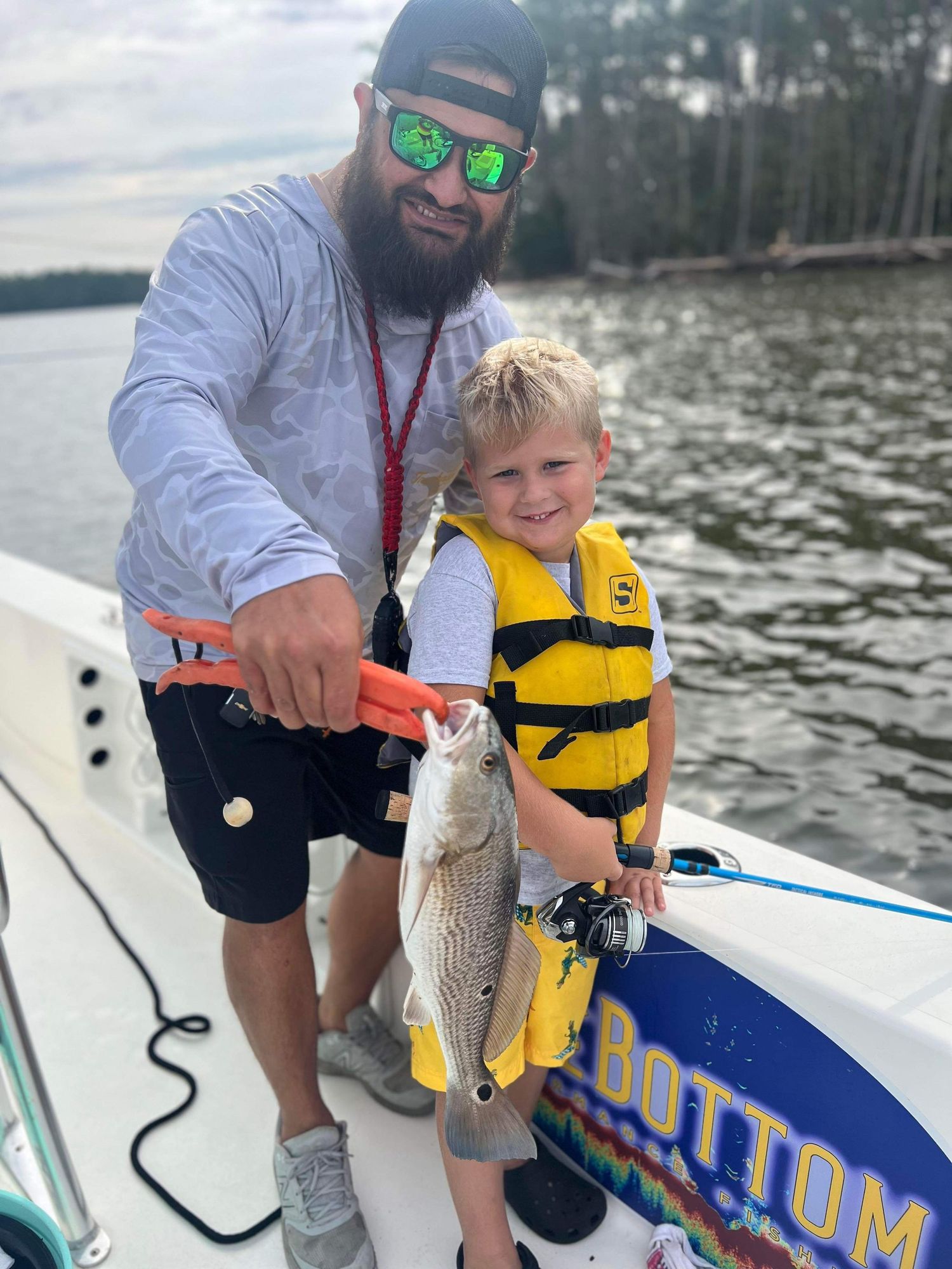 Two people showing off their catch on fishing boat near wooded shoreline.