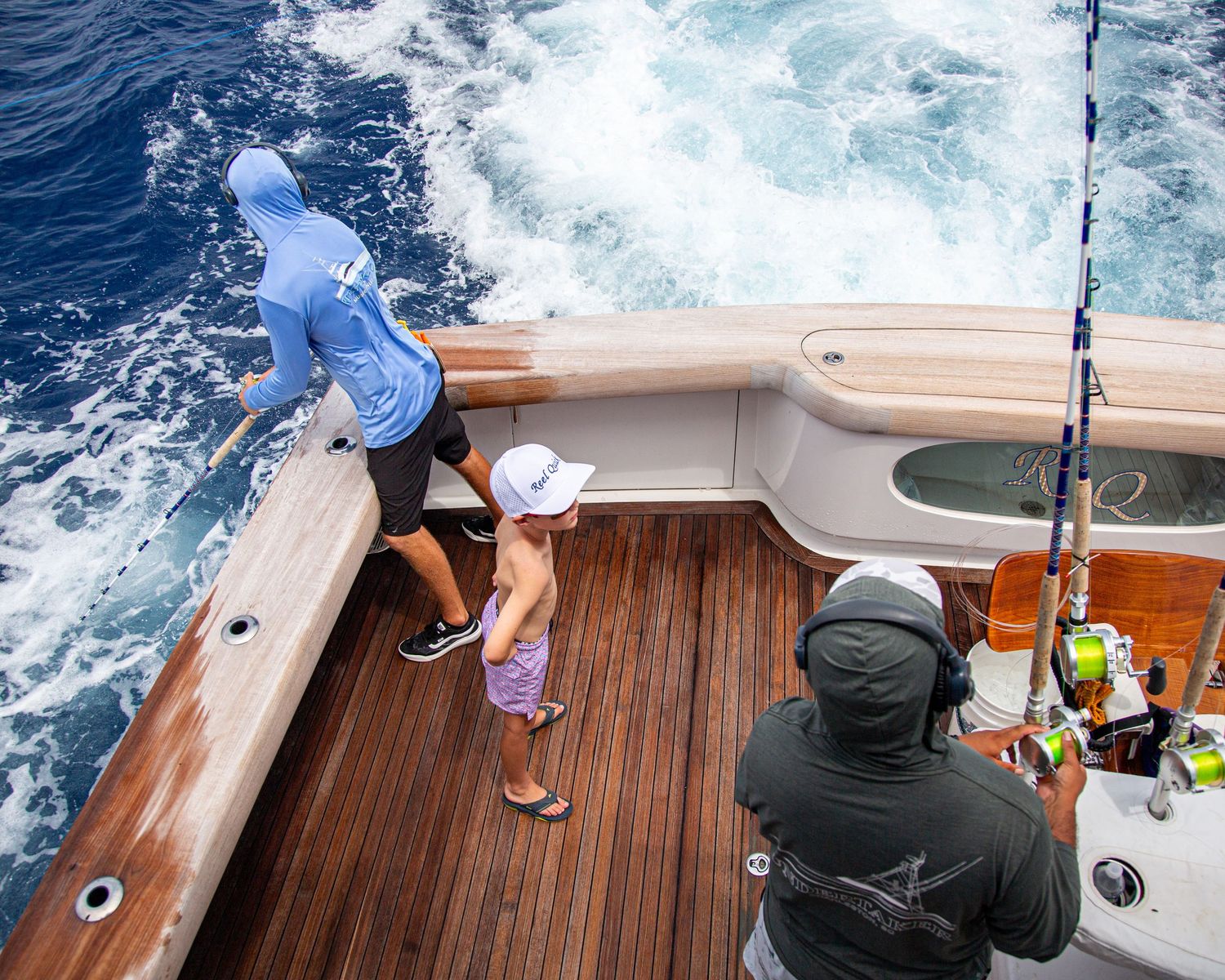 Anglers fishing from a sport fishing boat deck with trolling rods, blue ocean waves in background.