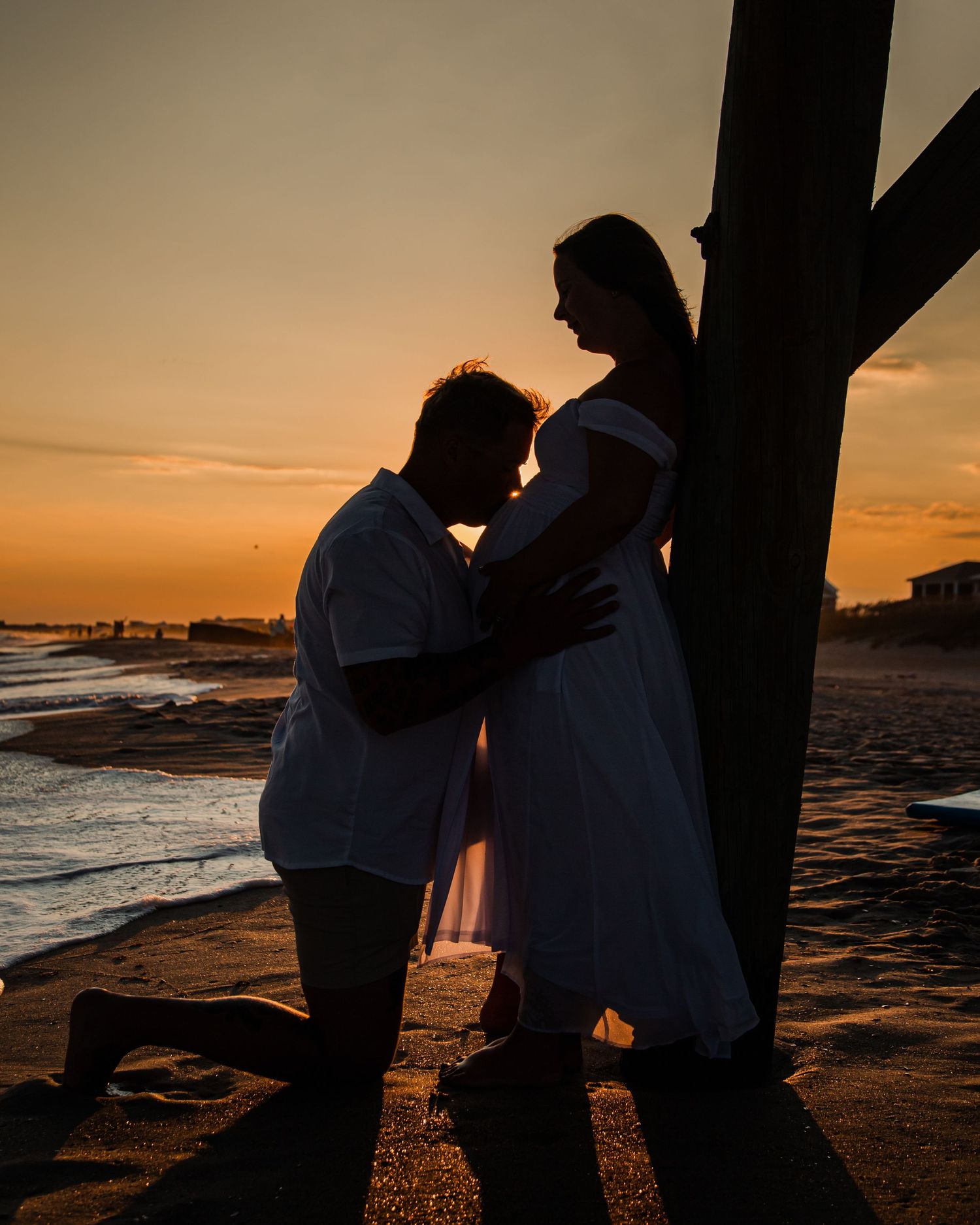 Silhouette of a couple at sunset on the beach, man kneeling before pregnant woman in white dress near a pier.