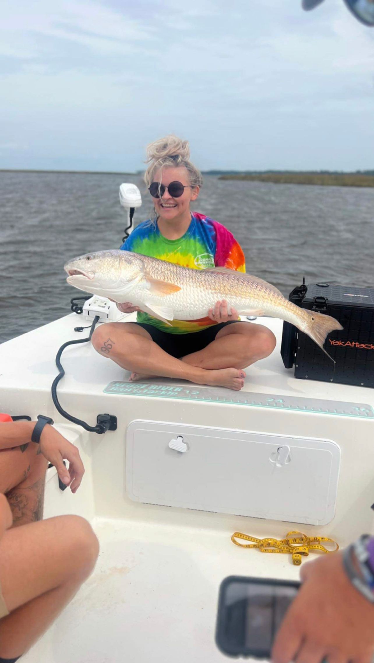 Angler wearing tie-dye shows off large redfish while sitting cross-legged on boat deck.