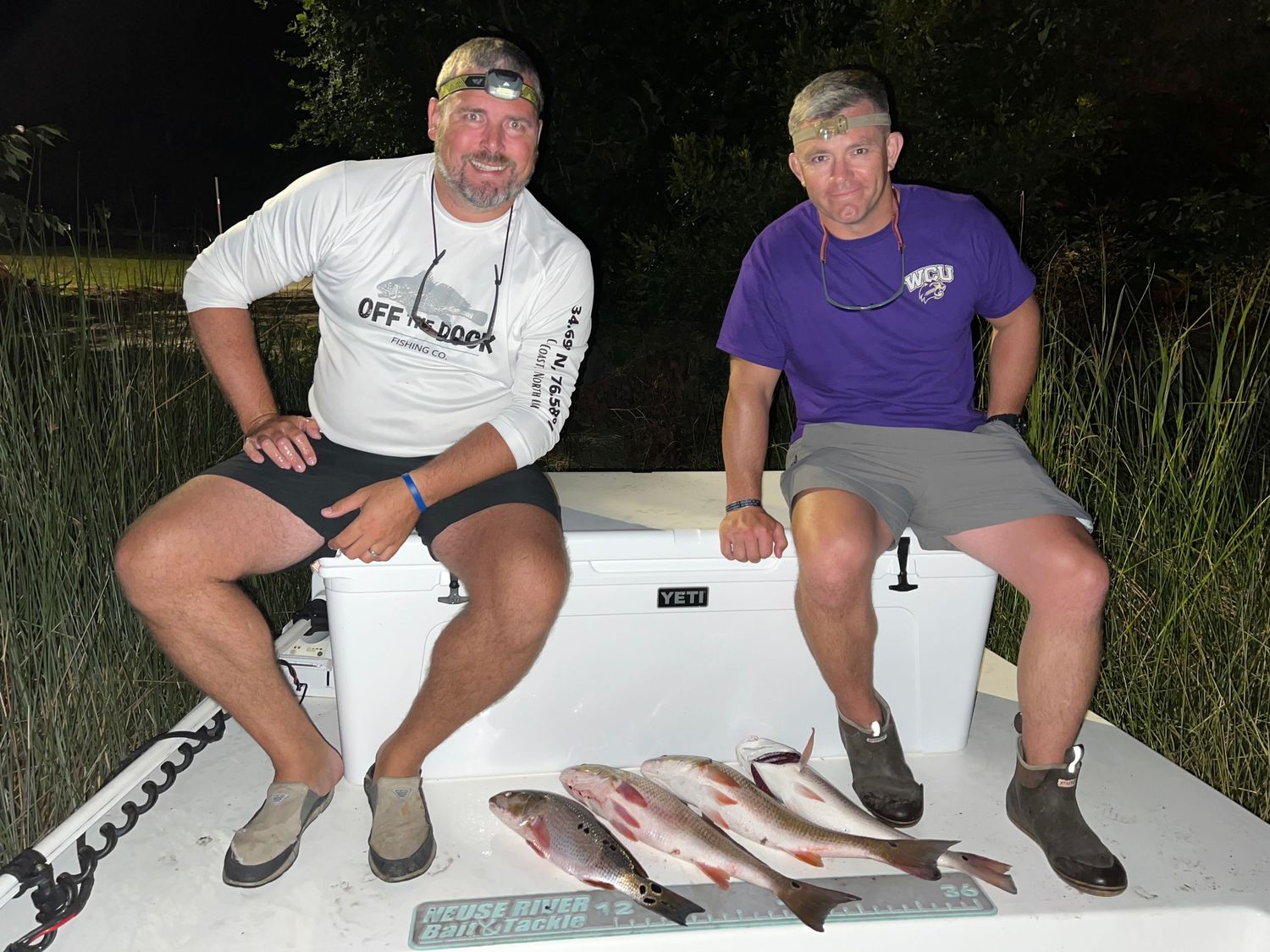 Two anglers display multiple fish catches while seated on white boat deck.
